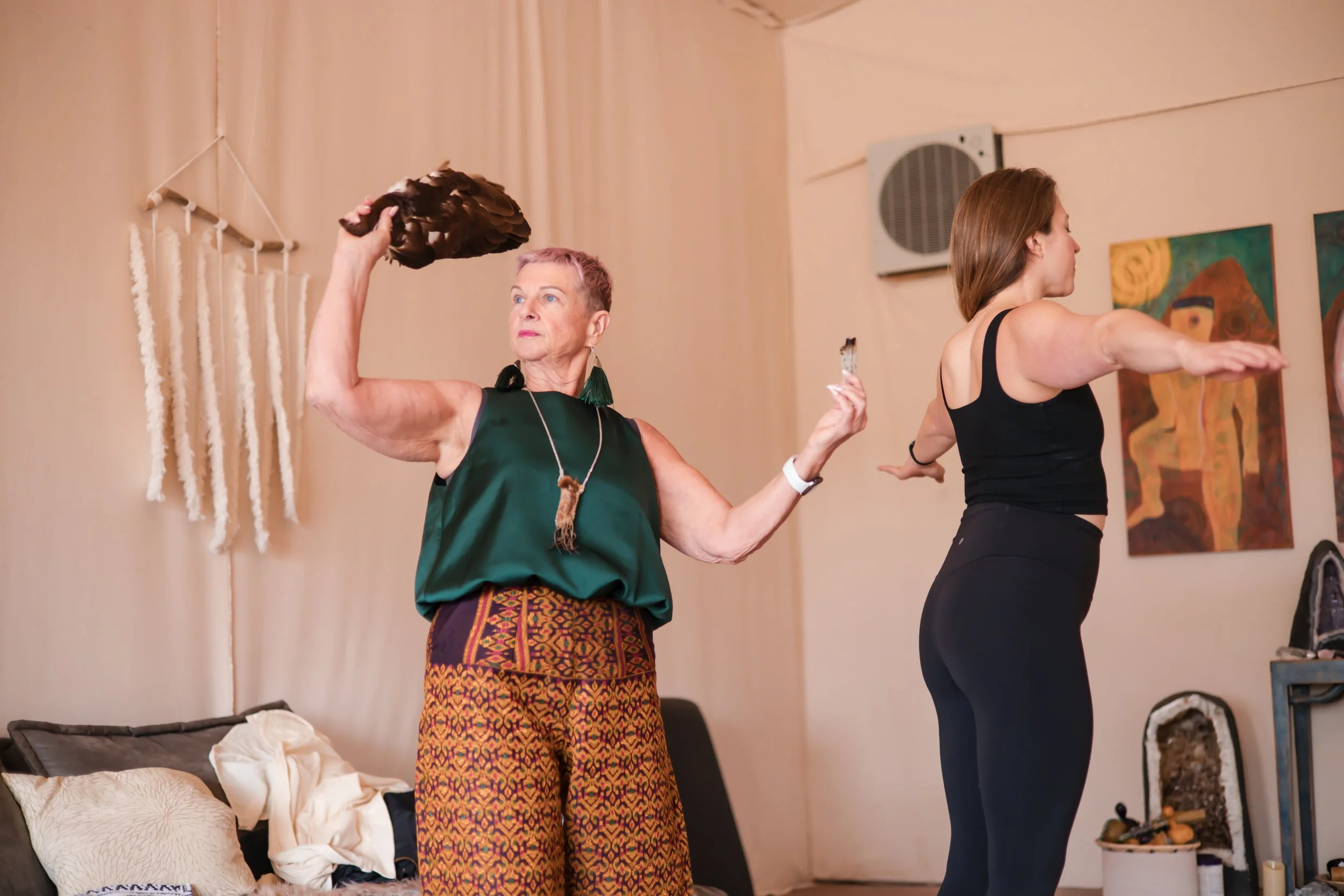 Two women in athletic clothing stretching and dancing in a room with light-colored walls and abstract art, one older woman with short purple hair holding a brown feathered fan, and a younger woman with long brown hair extending her arms