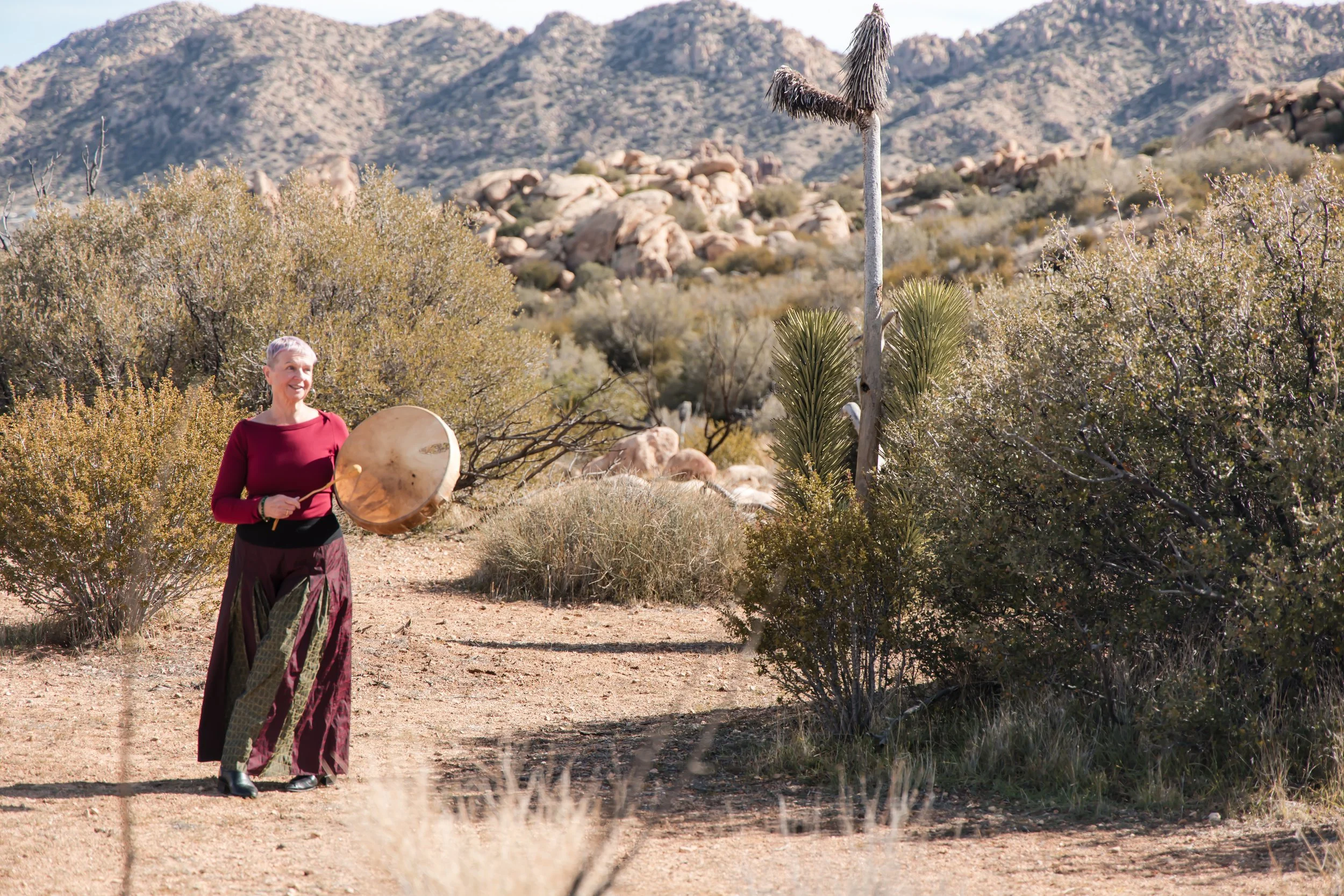 A woman in a red top and long skirt playing a gong on a dirt path in a desert landscape with shrubs, cacti, and rocky mountains in the background.