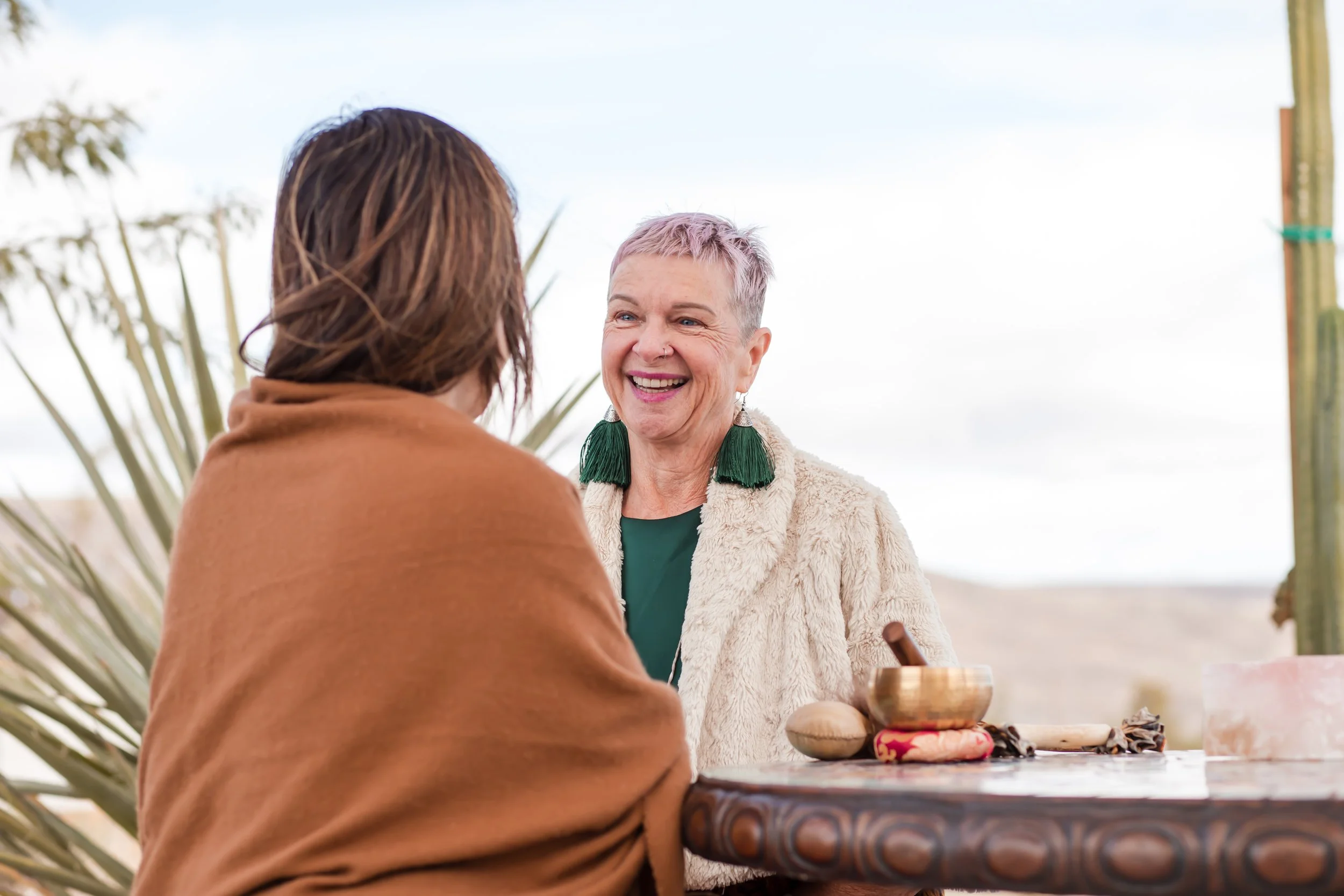 Two women smiling and talking outdoors at a table with a singing bowl, a stone, and a cloth, with desert plants and mountains in the background.