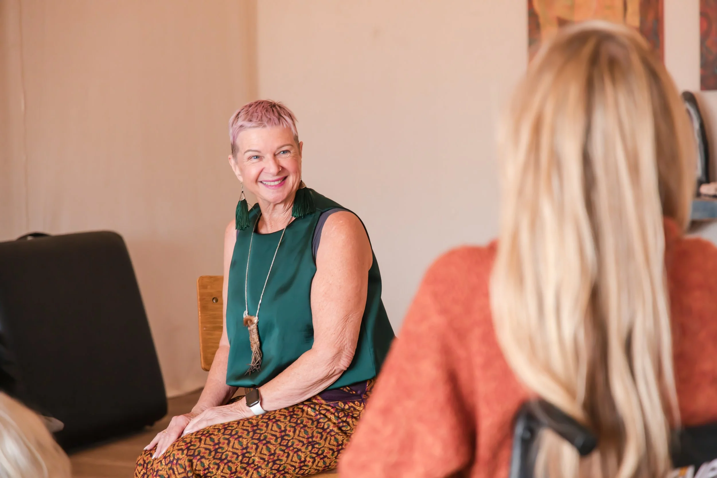Two women having a conversation, one with short pink hair and a colorful skirt, smiling, and the other with long blonde hair, wearing a rust-colored sweater, seen from behind.