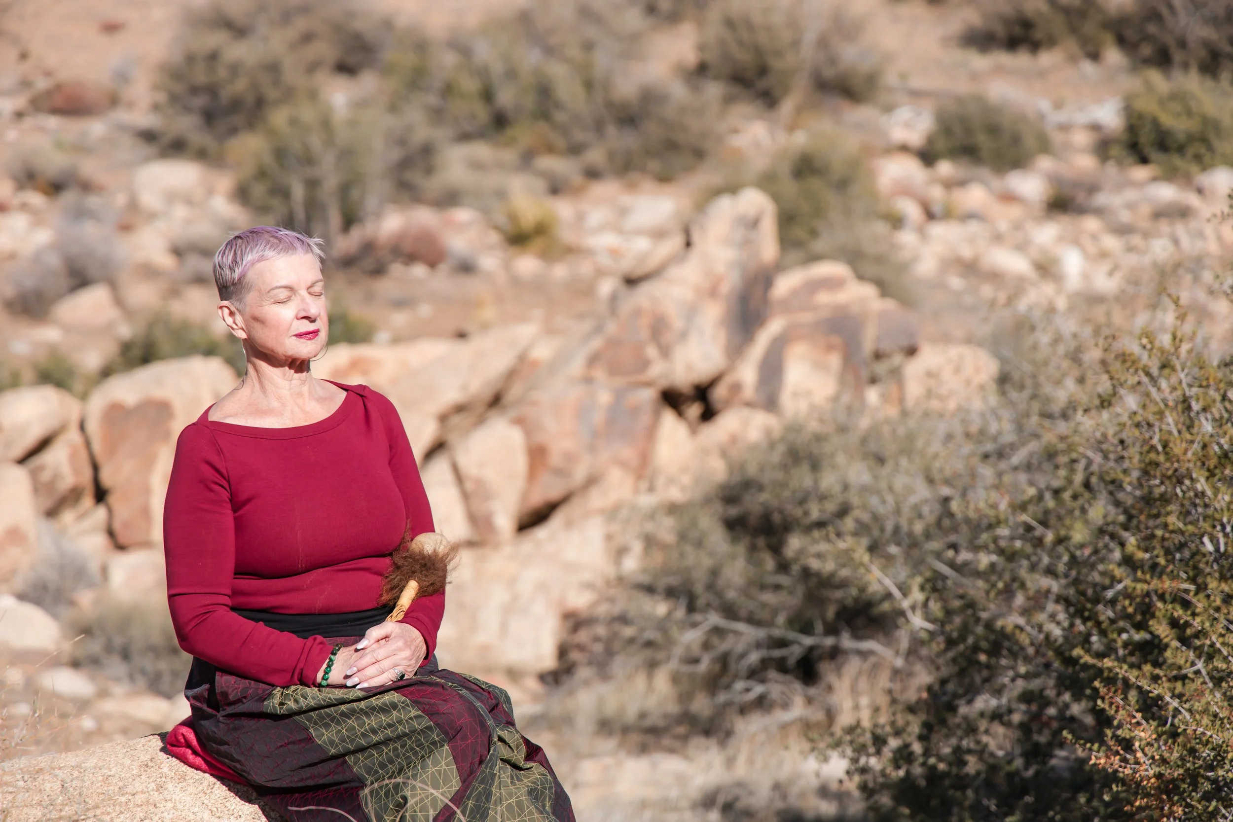 An elderly woman with short gray hair, wearing a red long-sleeve top, sitting outdoors on a rock with her eyes closed and meditating in a rocky desert landscape.