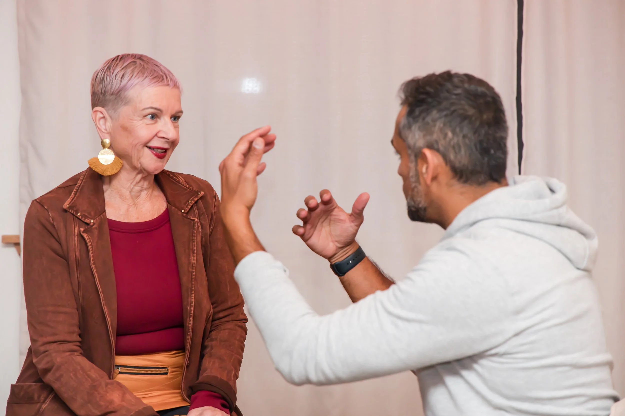 An older woman with short pink hair and large yellow earrings is smiling and talking to a man with gray hair and a beard, who is gesturing with his hands during a conversation in front of a plain light-colored background.