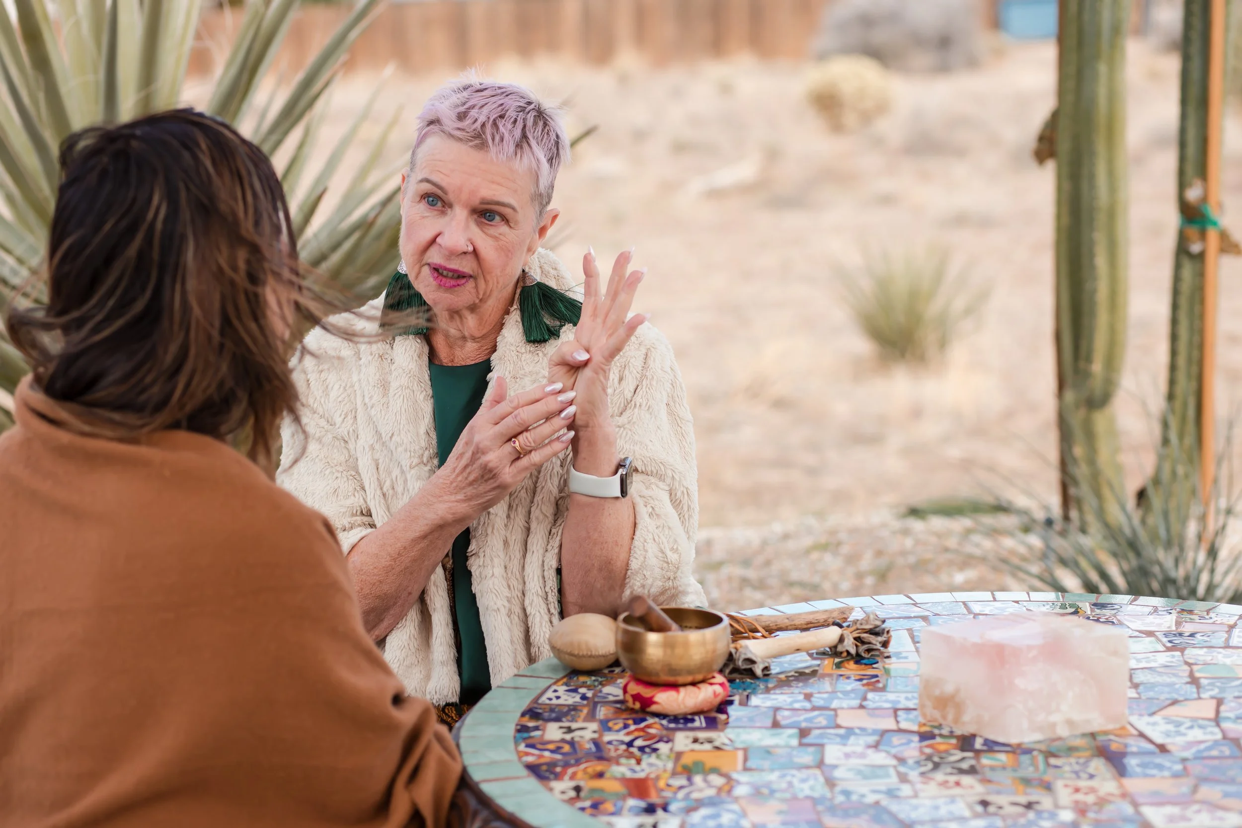 Two women engaged in a tarot card reading outdoors, with desert plants like agave and cacti in the background. One woman has short grey hair and is speaking, while the other has long brown hair. The table between them has tarot cards, a pink salt lamp, and various small objects.