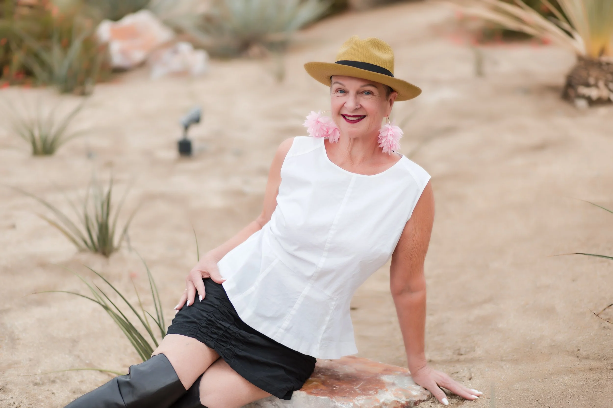 A smiling woman sitting on a rock in a desert-like setting with succulents, wearing a straw hat, pink feather earrings, a white sleeveless top, black shorts, and black boots.