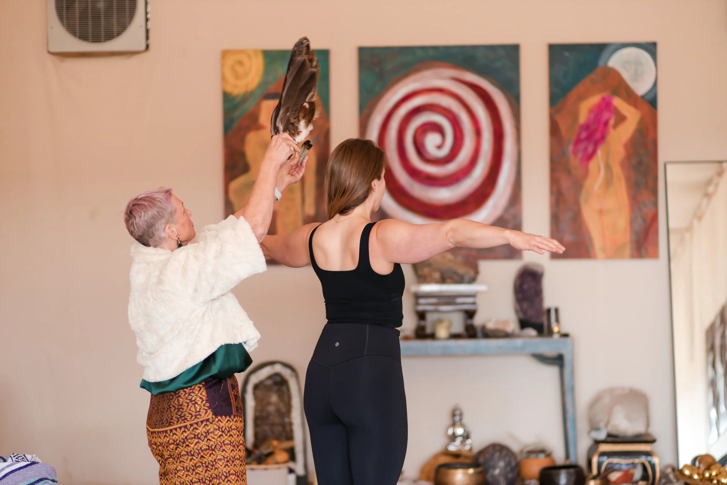 An older woman with white hair and colorful patterned skirt stands beside a woman in a black tank top and leggings, performing a yoga pose with her arms extended. The older woman raises her arm holding a large bird, possibly a hawk or eagle, in an art-filled indoor space with abstract paintings on the wall.