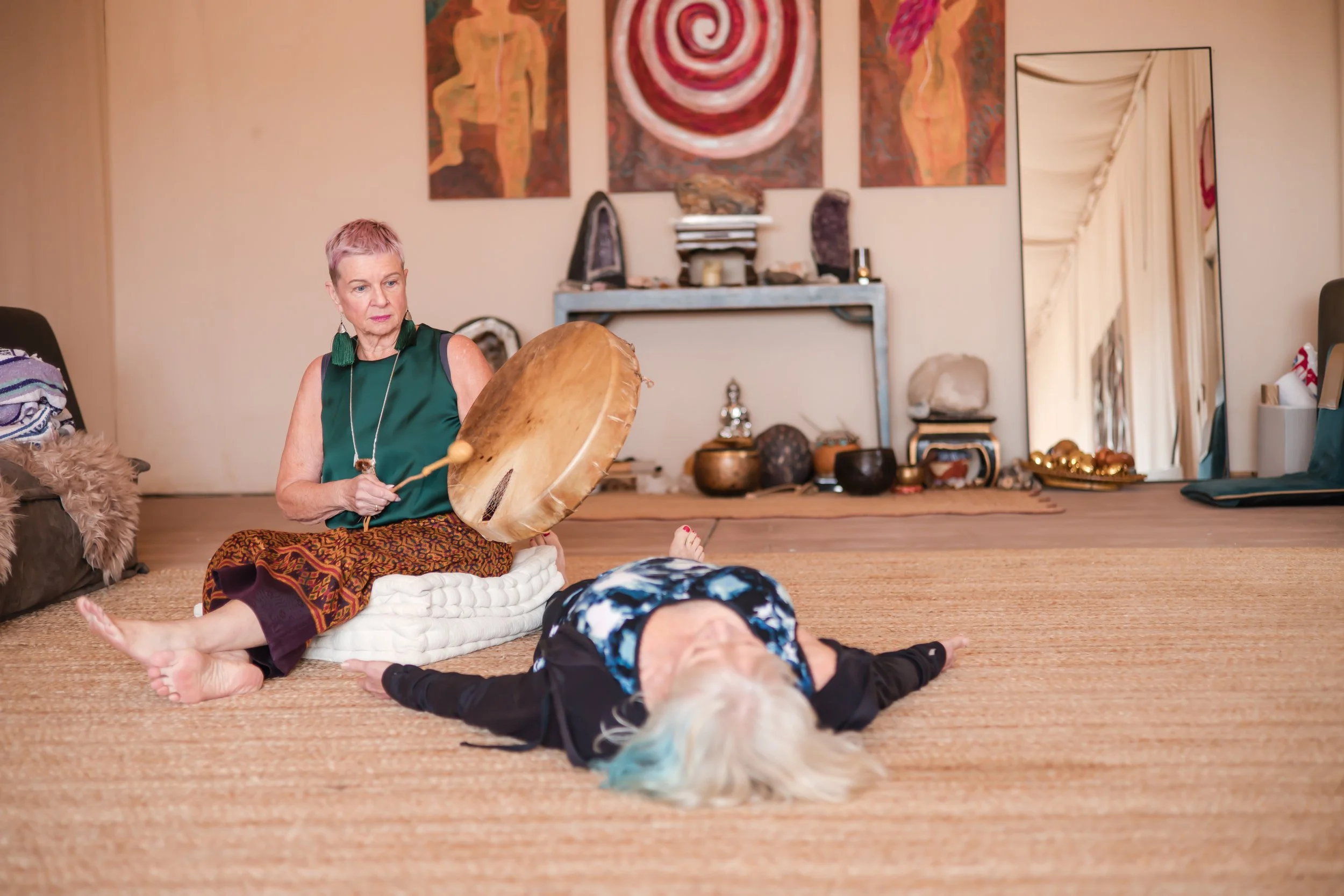 An older woman lying on the floor with platinum blonde hair with hints of blue, eyes closed, in a yoga pose, wearing a black and blue patterned top. Next to her, a woman with short pink hair, wearing a green top and patterned skirt, sitting on the floor playing a large drum with a mallet, in a room decorated with artwork, crystals, and spiritual objects.