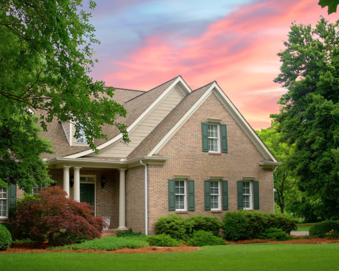 A brick house with dark green shutters, a covered front porch, surrounded by green trees and bushes, under a pink and purple sunset sky.