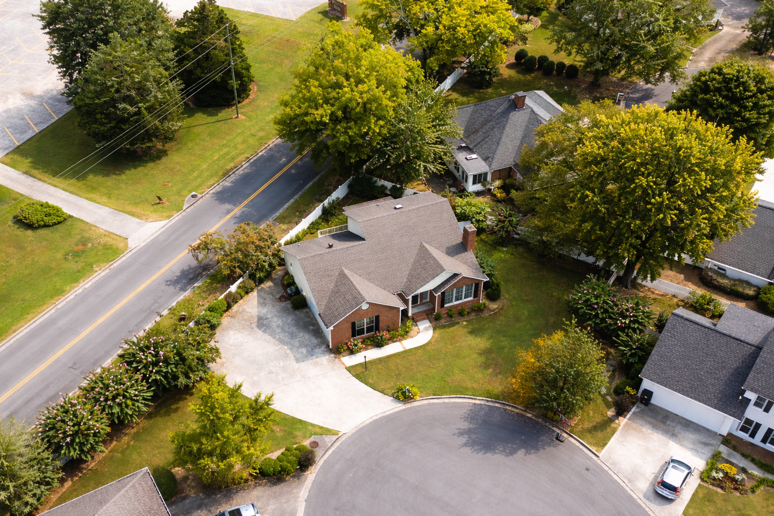 An aerial view of a residential neighborhood featuring several houses, driveways, and lush green trees along a quiet street.