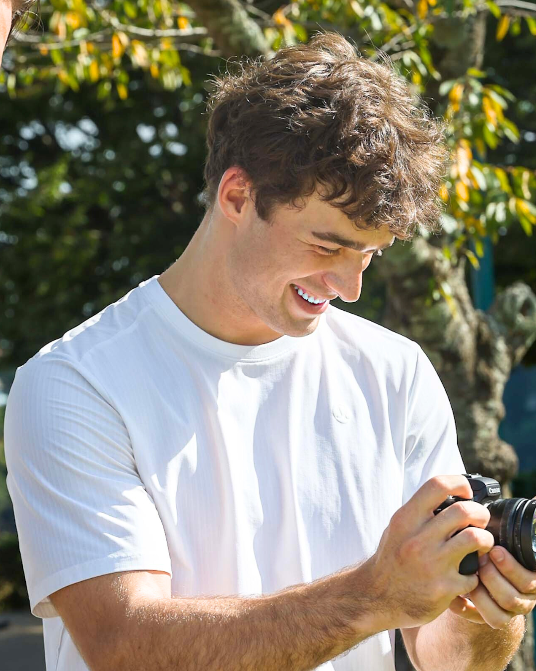 A young man with curly brown hair, wearing a white shirt, is outdoors smiling while looking at a camera he is holding.