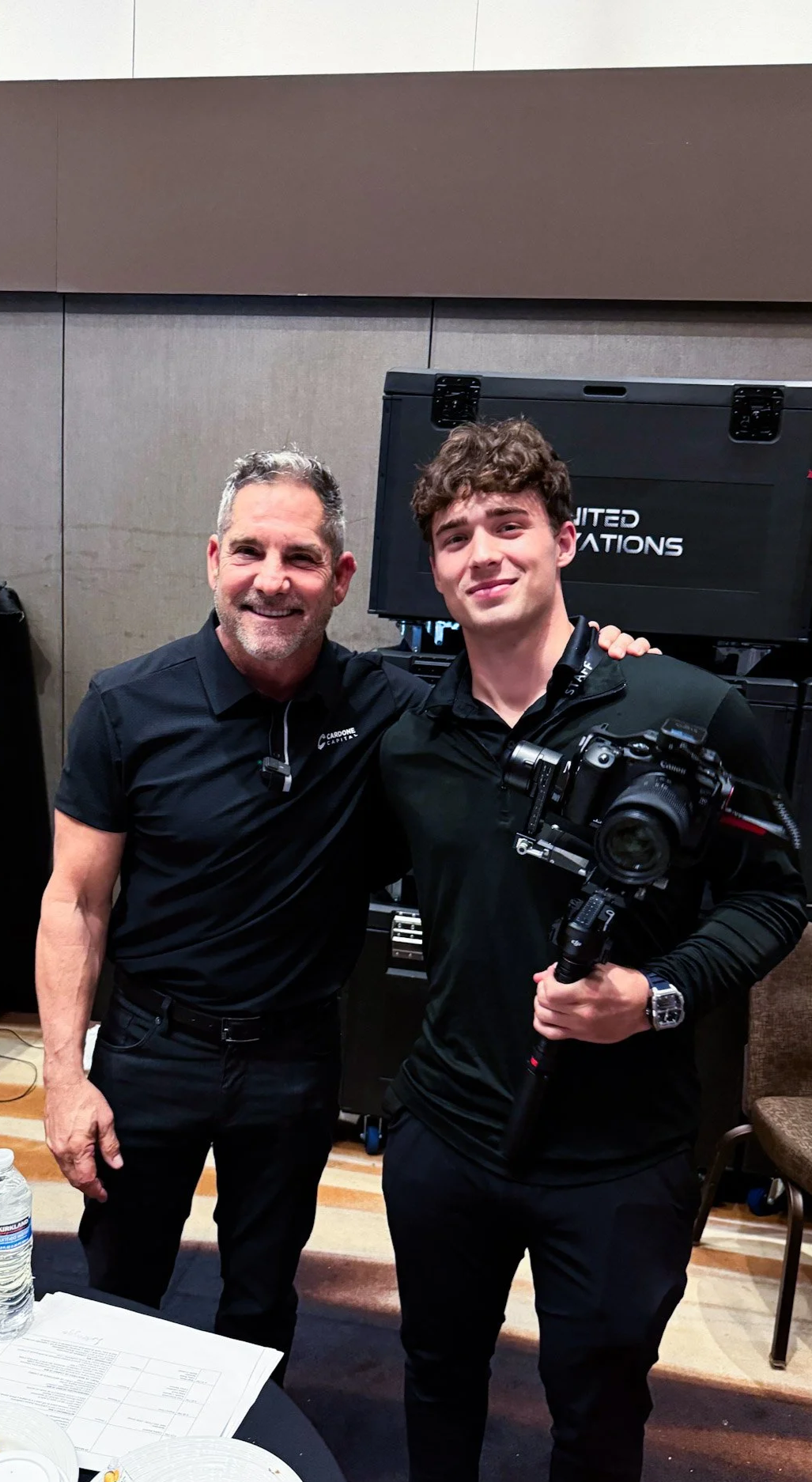 Two men standing close together, smiling at the camera, in an indoor setting. The man on the left has gray hair and a beard, wearing a black shirt with a logo. The man on the right has curly brown hair, is clean-shaven, wearing a black shirt, and holding a professional camera with a stabilizer. They are in front of a black equipment case with the printed text "United Nations".