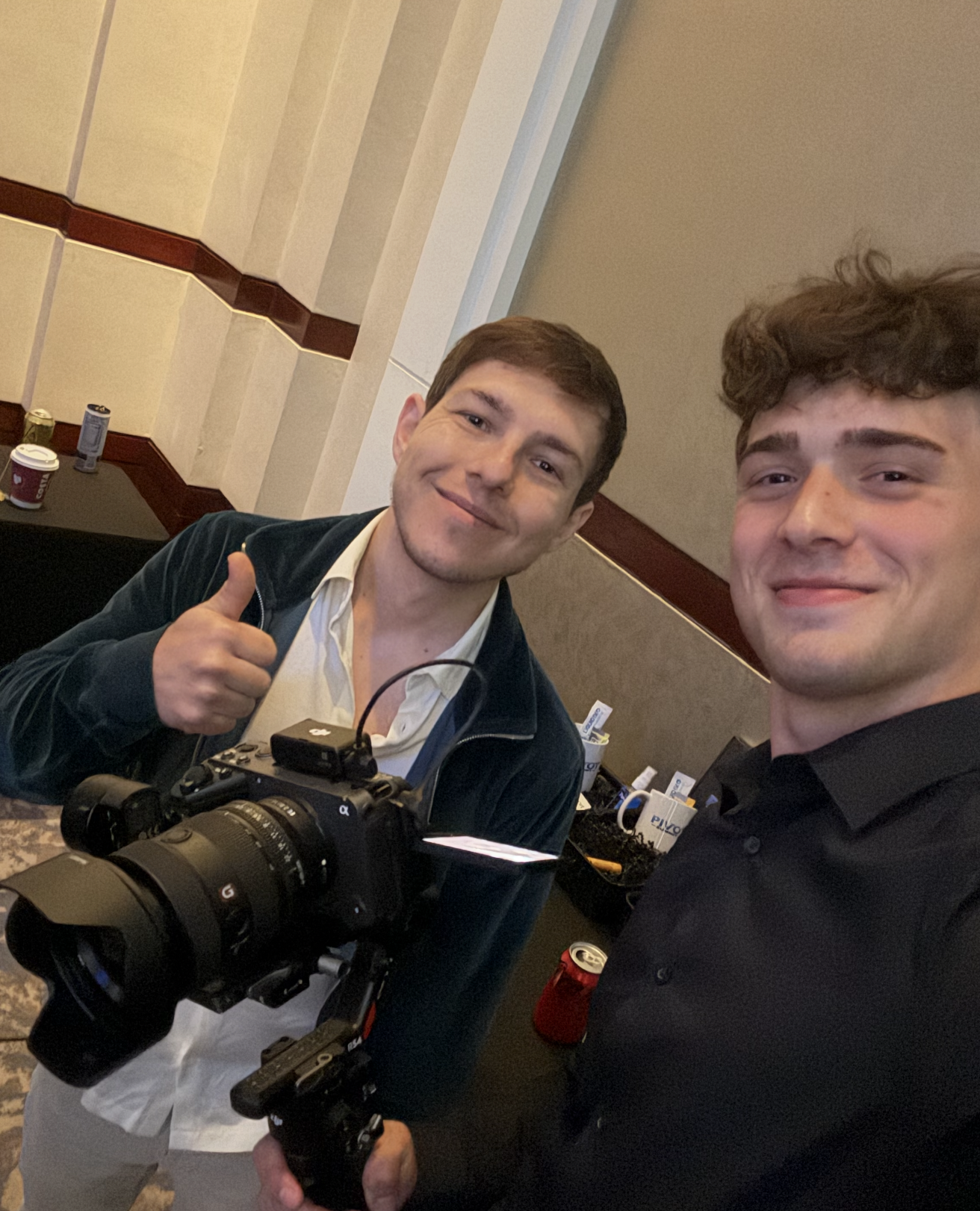 Two young men smiling and taking a selfie at an indoor event. One is holding a professional camera with a large lens.