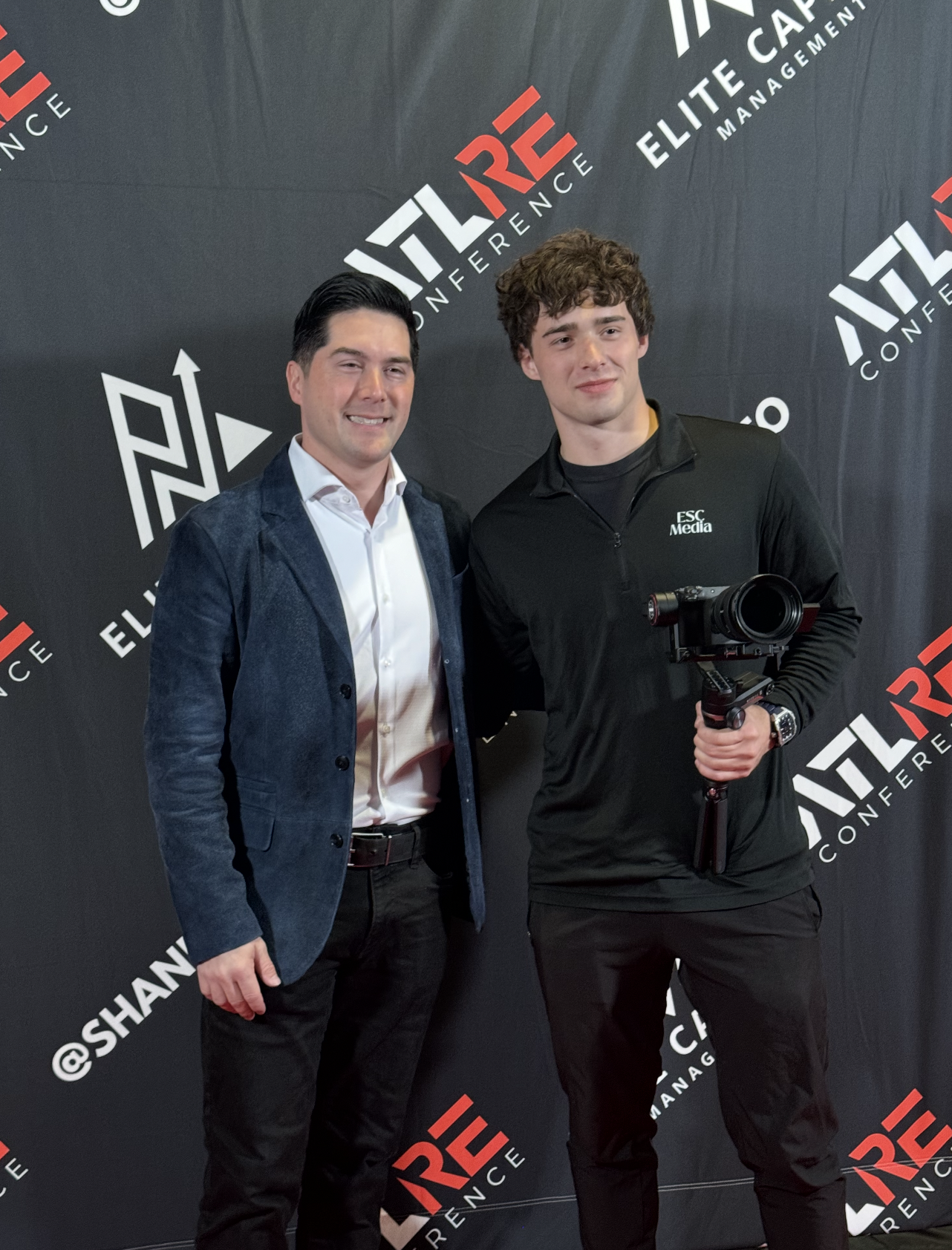 Two men standing in front of a black backdrop with white and red text, smiling at the camera. One man is holding a camera stabilizer. The backdrop has logos and words related to a conference.