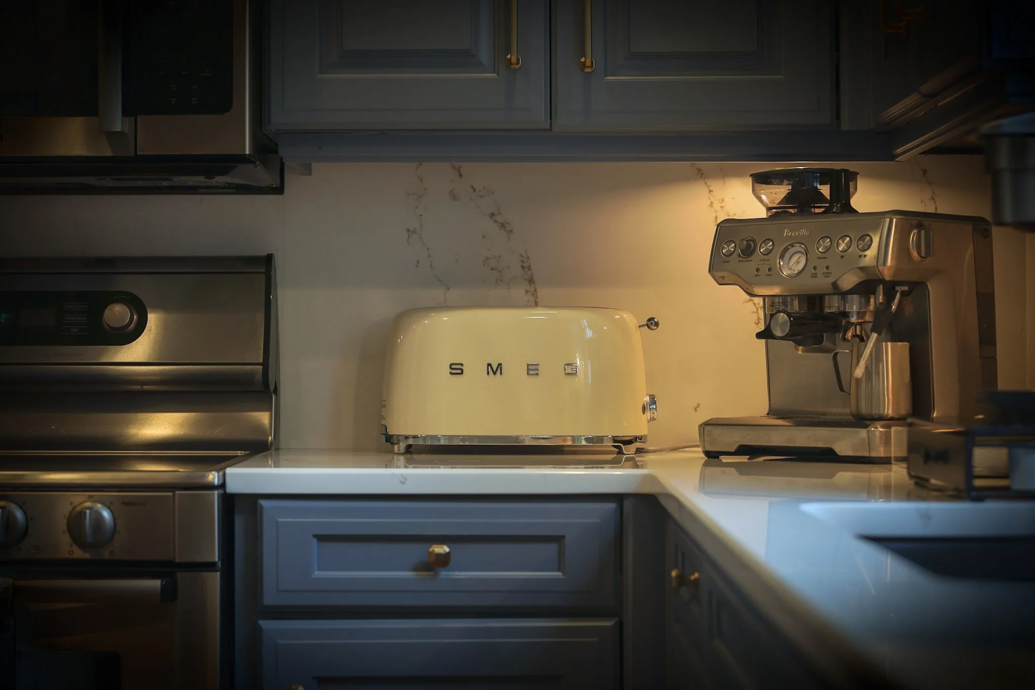 Kitchen countertop with a vintage Smeg toaster between an oven and a coffee machine, with gray cabinets and marble backsplash.