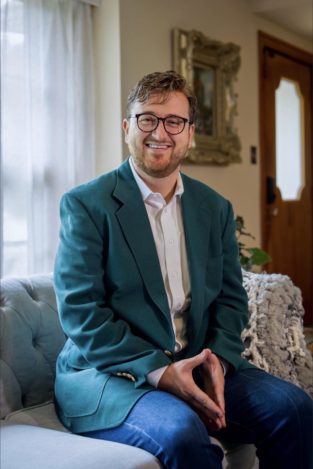 A man with brown hair, glasses, and a beard, wearing a teal blazer and white shirt, sitting on a couch and smiling inside a cozy, well-lit room with a wooden door and decorative mirror in the background.