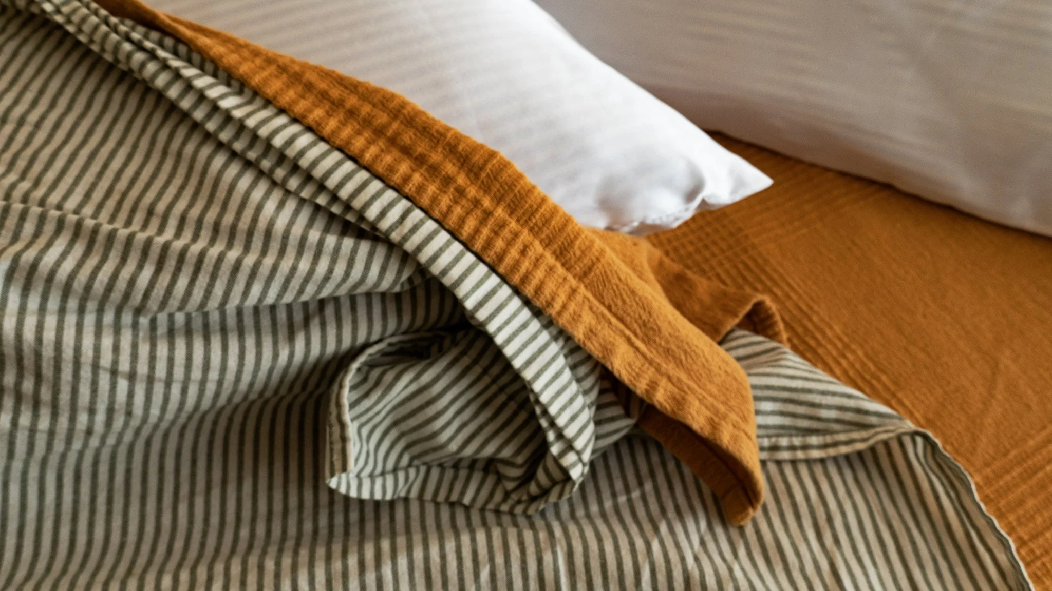 Close-up of a bed with a white pillow, a brown blanket, and a striped bedspread with a fold showing orange and beige colors.