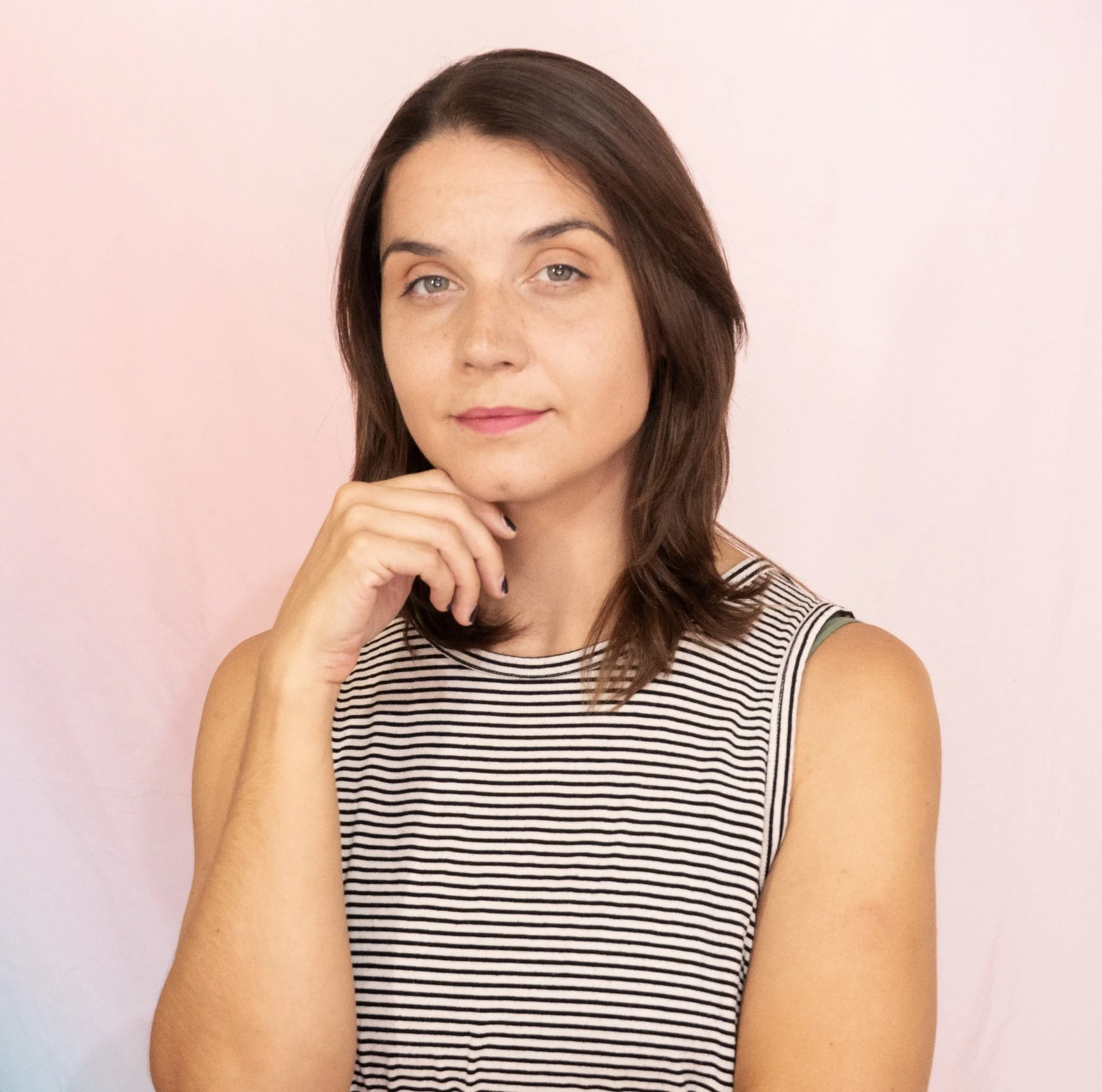 A woman with shoulder-length brown hair, light skin, and blue eyes, wearing a black and white striped sleeveless top, resting her chin on her hand, standing against a pale pink wall.