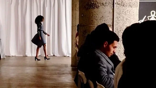 A woman in a gray dress and high heels walking on a stage with a white curtain backdrop, while three people sit in the foreground, partially visible. The setting appears to be an event or performance space.