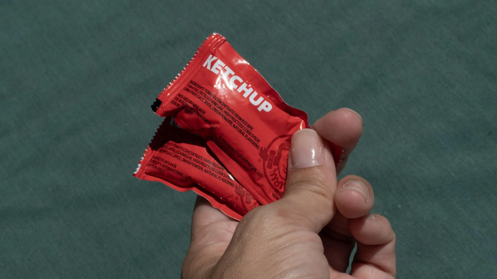 Close-up of hand holding a red packet of ketchup against a teal background.
