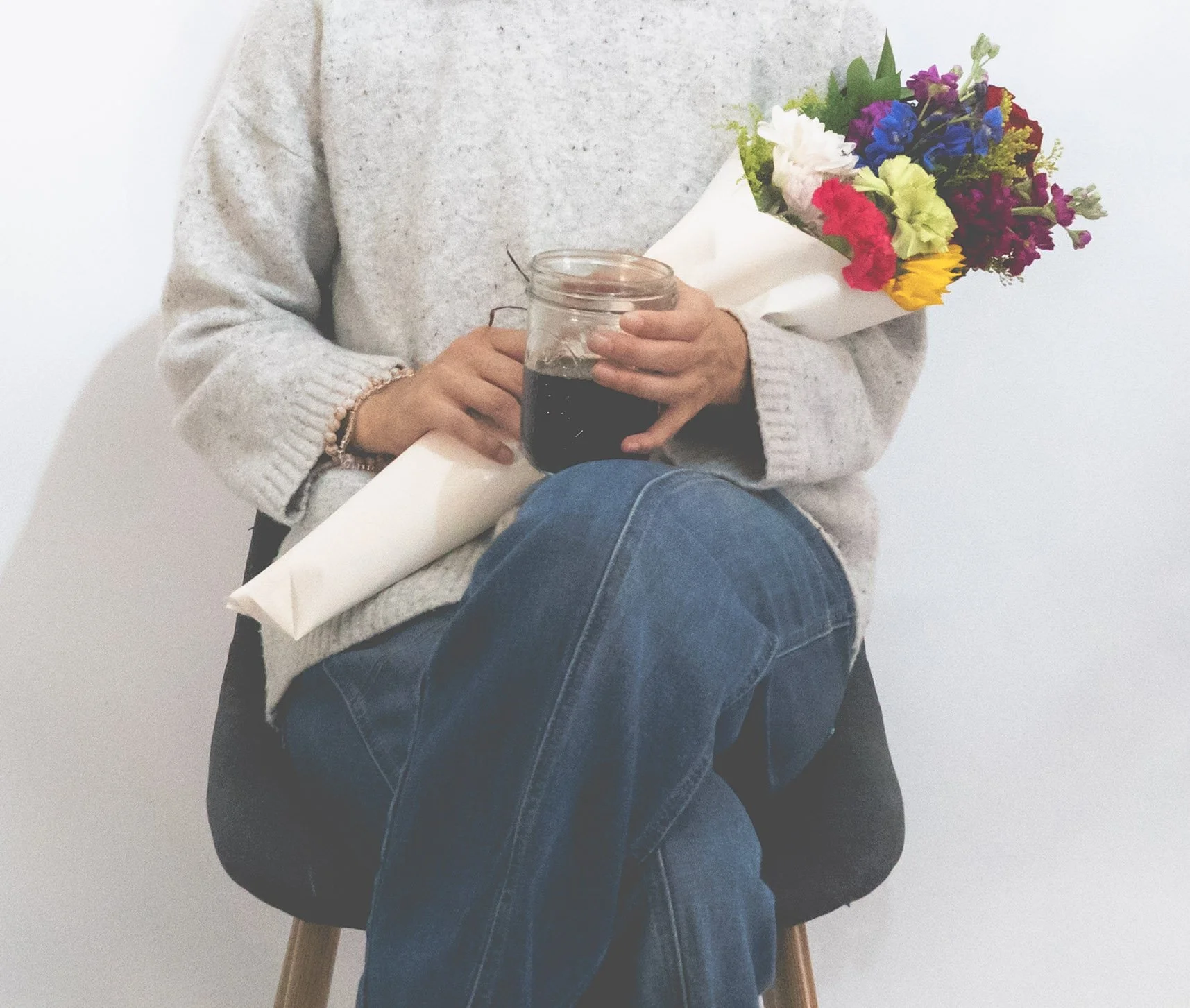 Person holding a bouquet of colorful flowers and a jar of dark liquid, sitting on a chair against a plain white wall.