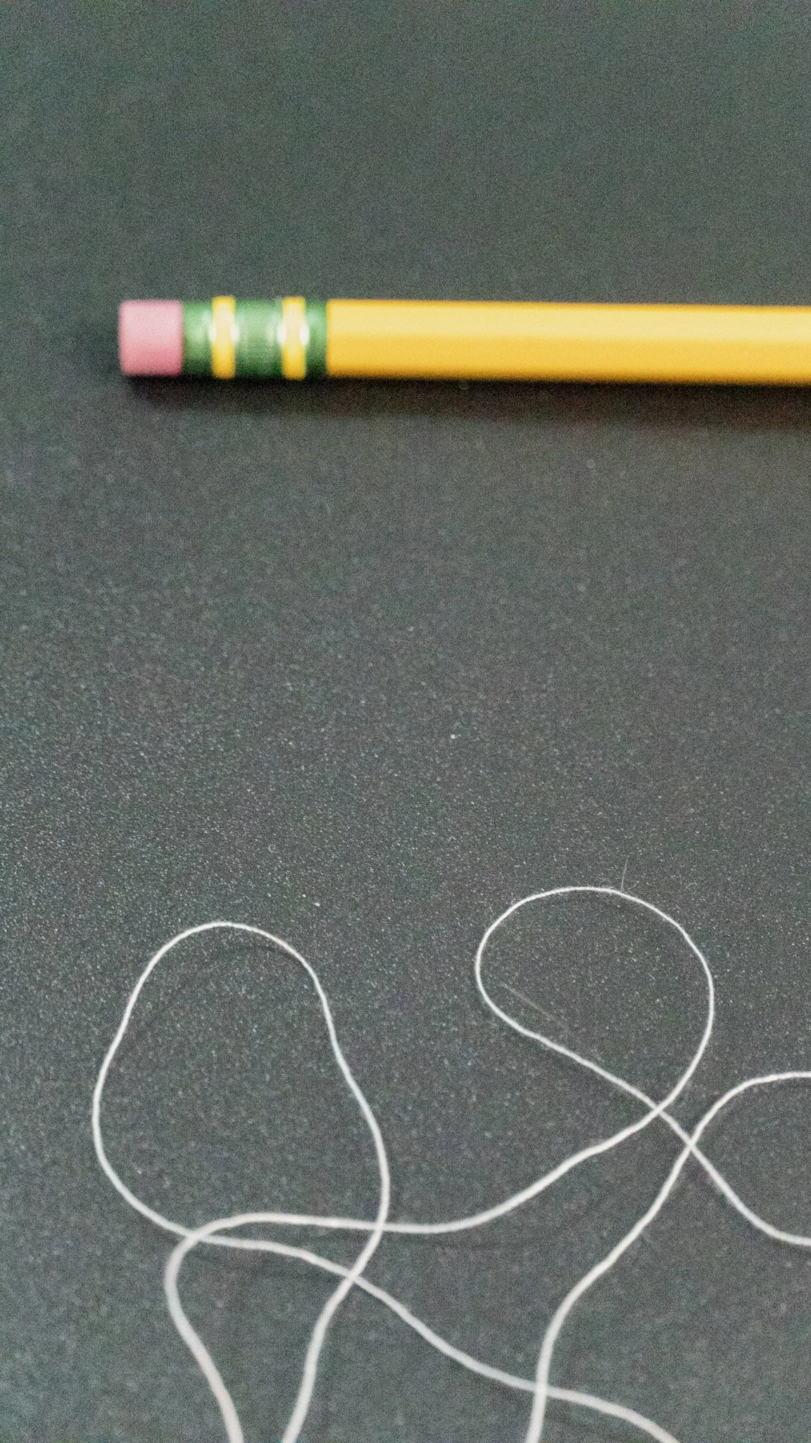 A yellow pencil with a pink eraser and green and yellow rings is lying on a dark surface. Several white loops of thread are also visible on the surface.