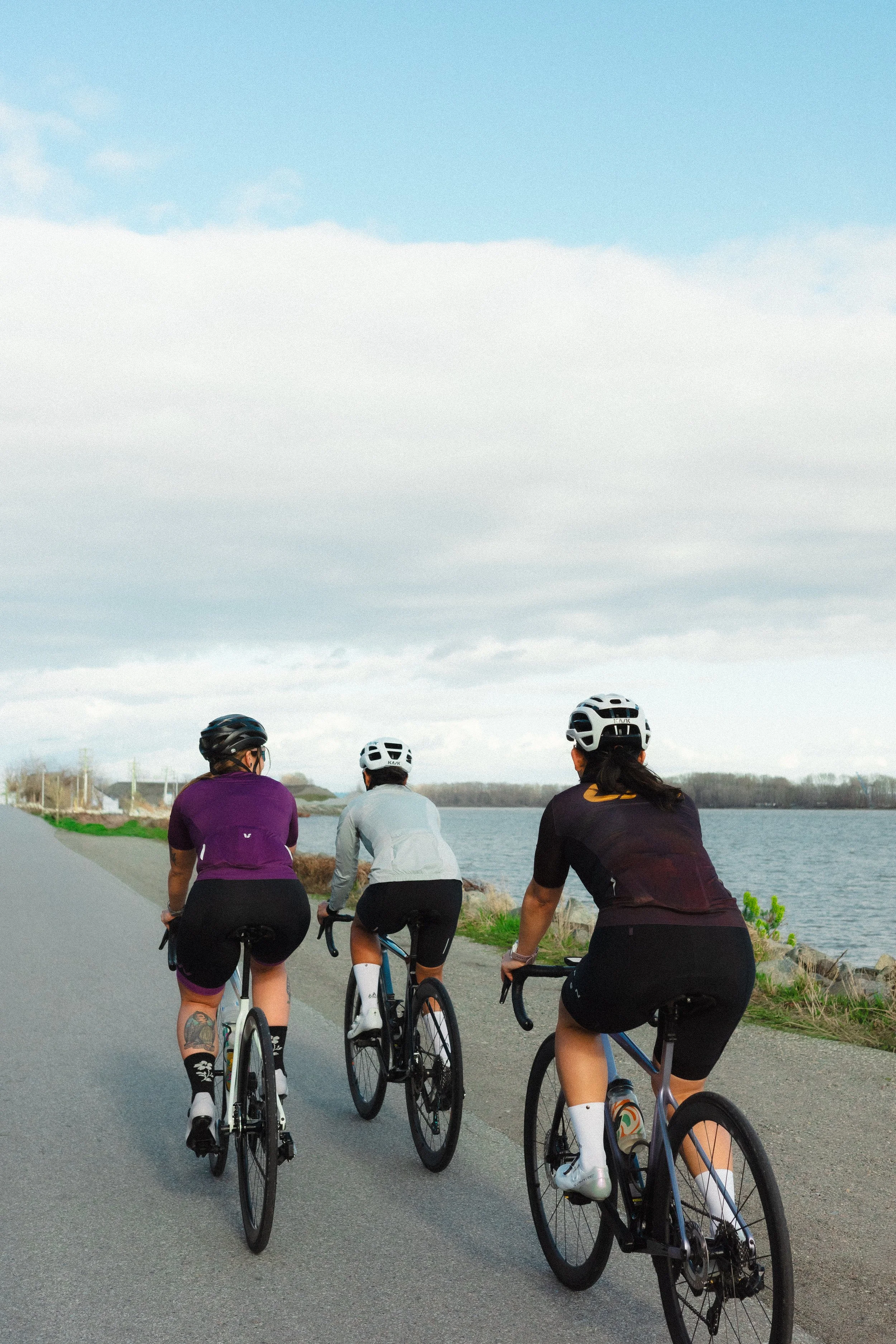 Three women wearing helmets riding bicycles along a shoreline on a paved path with water on one side and a cloudy sky above.