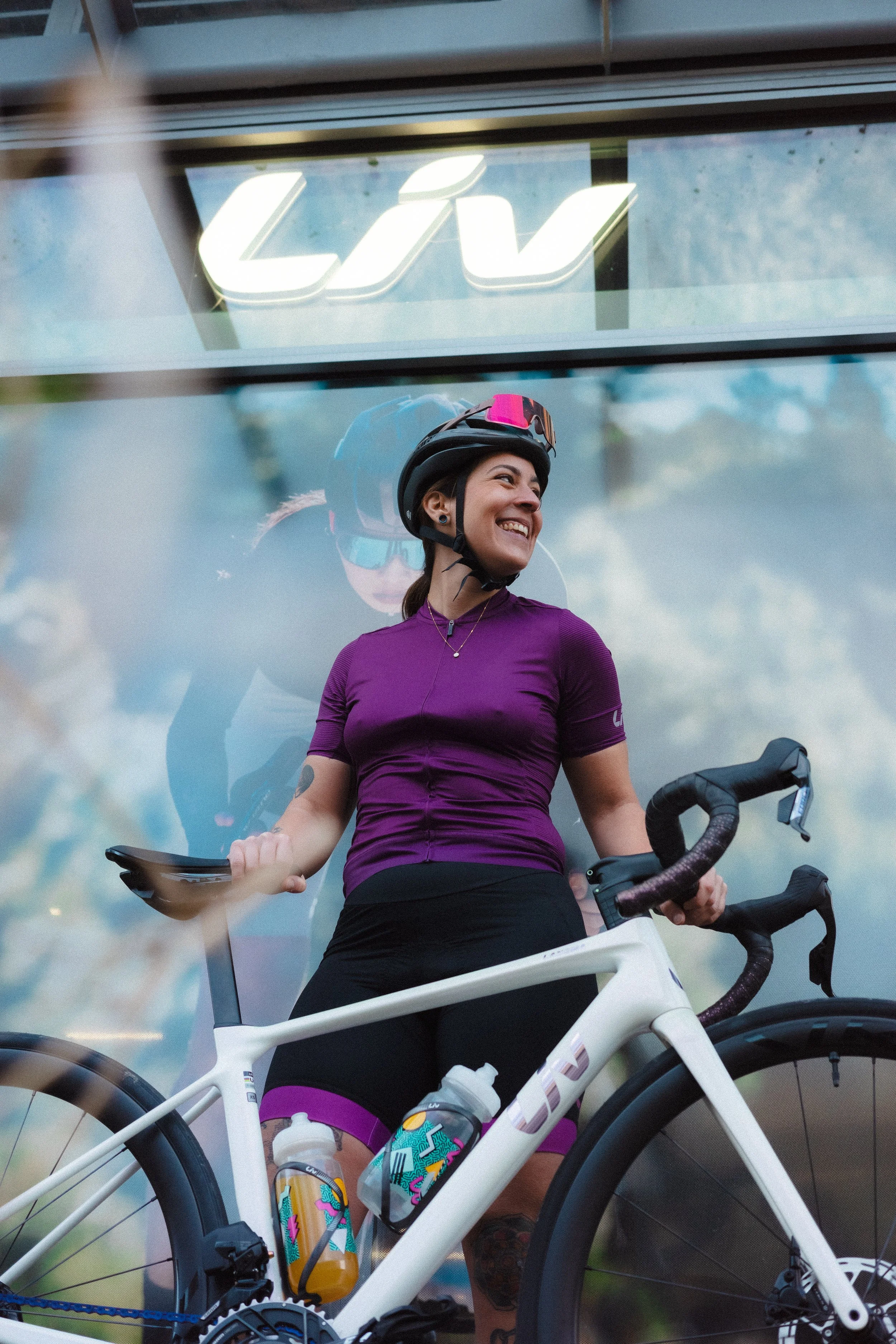 A woman in cycling gear, including a purple shirt and helmet, standing next to a white road bicycle with water bottles, smiling and looking to the side.