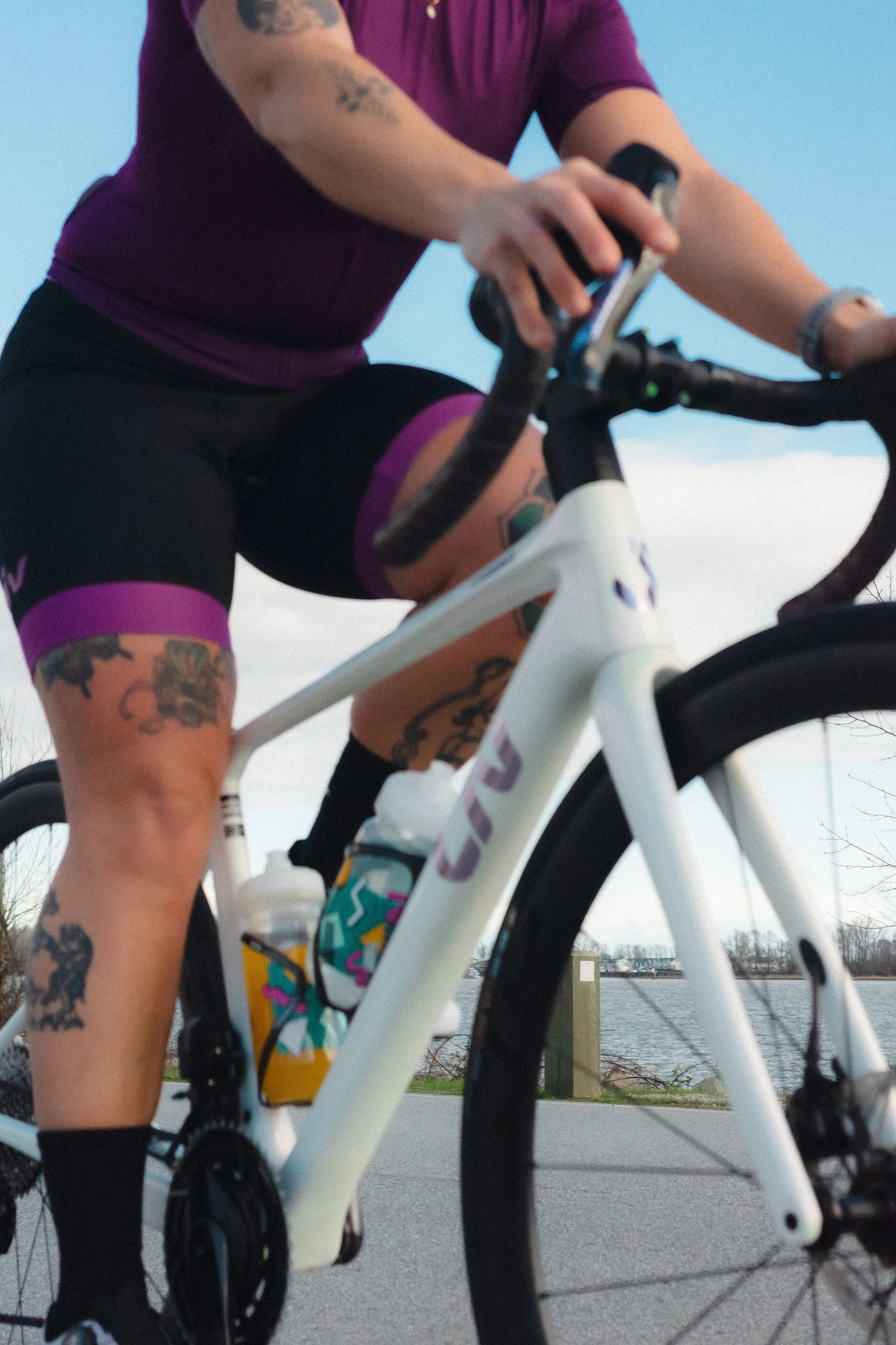 Close-up view of a person riding a white bicycle outdoors near a body of water. The person is wearing a purple short-sleeve shirt, black and purple cycling shorts, and black socks.