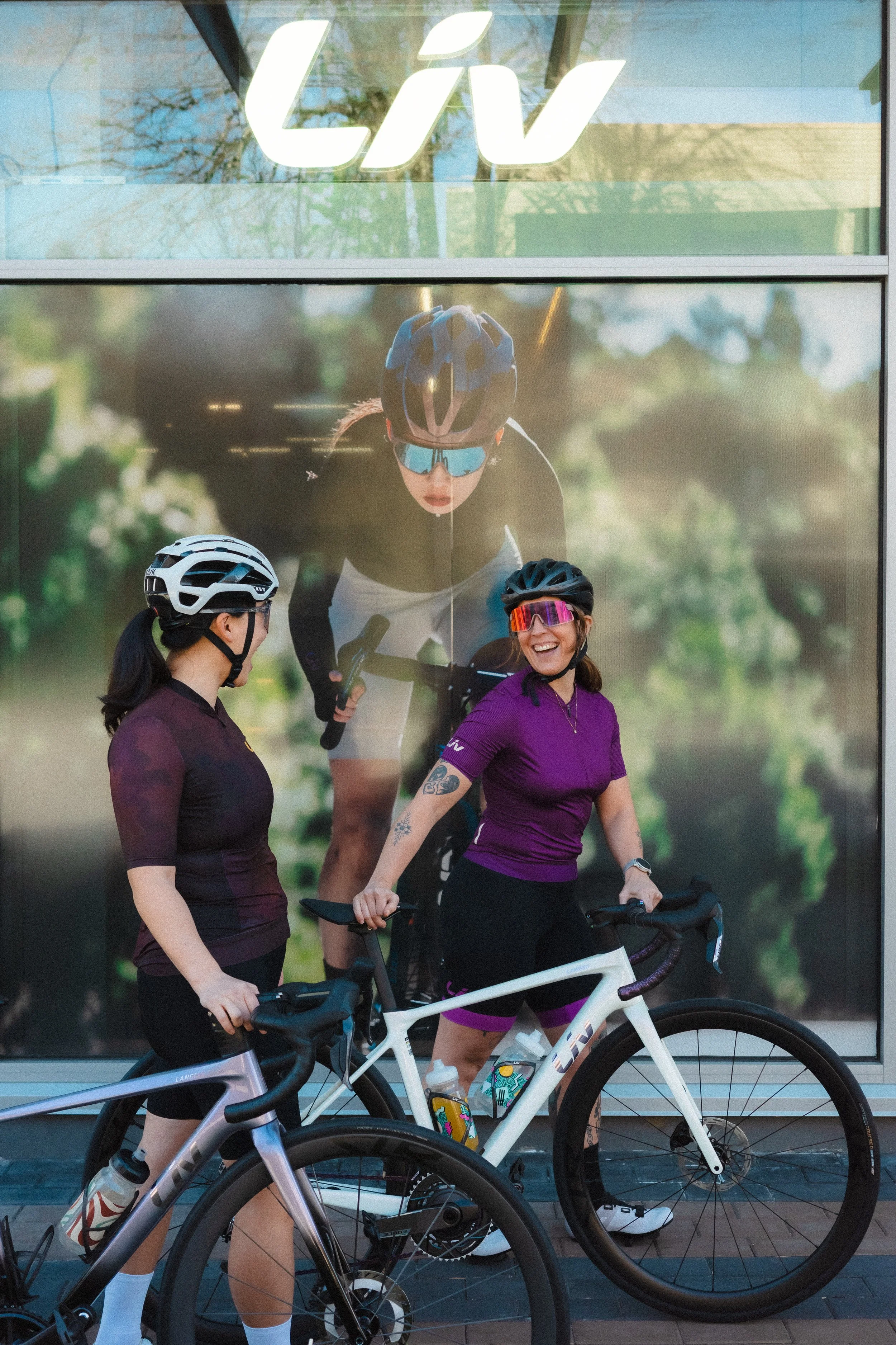 Two women in cycling gear standing with their bikes in front of a large, smiling cyclist poster at a LIV store.