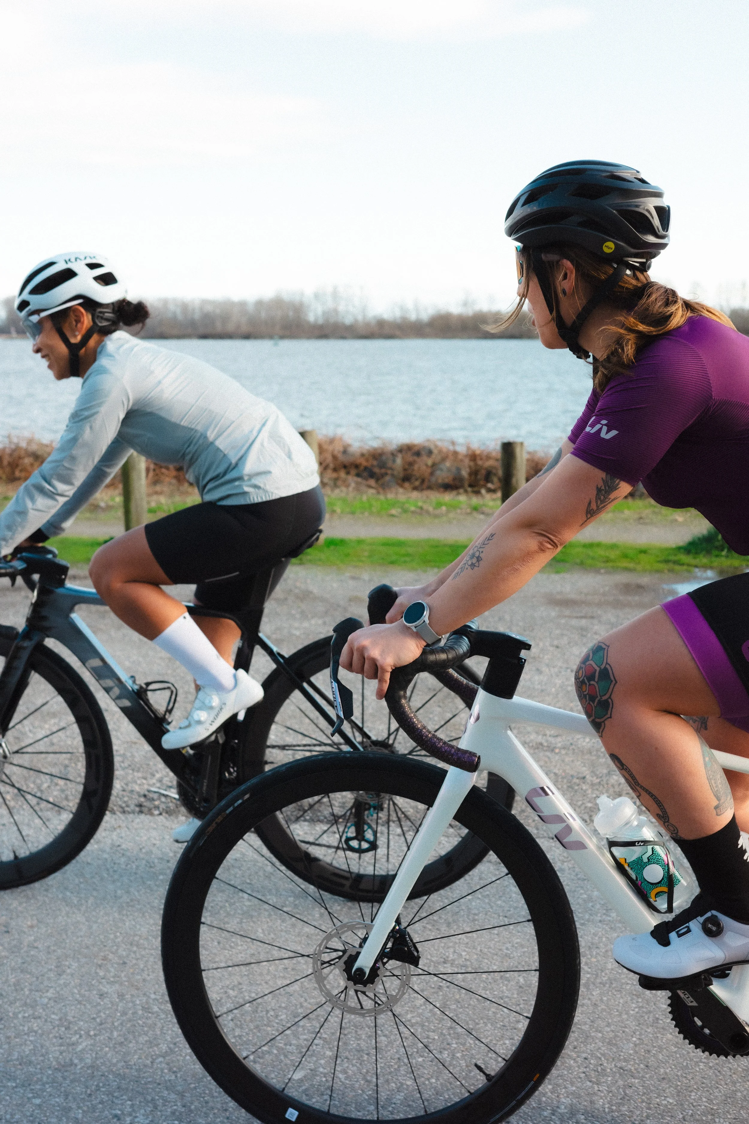 Two women riding bicycles outdoors near a body of water, one wearing a white helmet and the other wearing a black helmet, with a cloudy sky above.