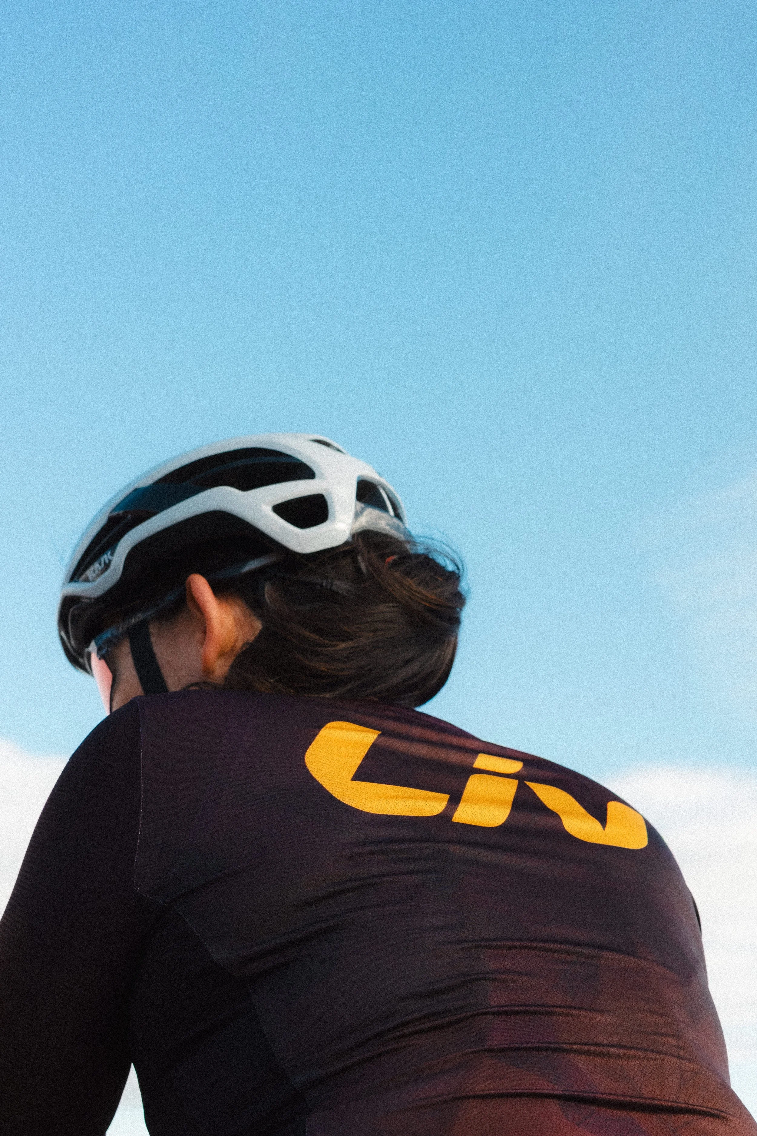 A woman wearing a bicycle helmet and a sports jersey with the logo 'CIN' on the back, looking away against a clear blue sky.