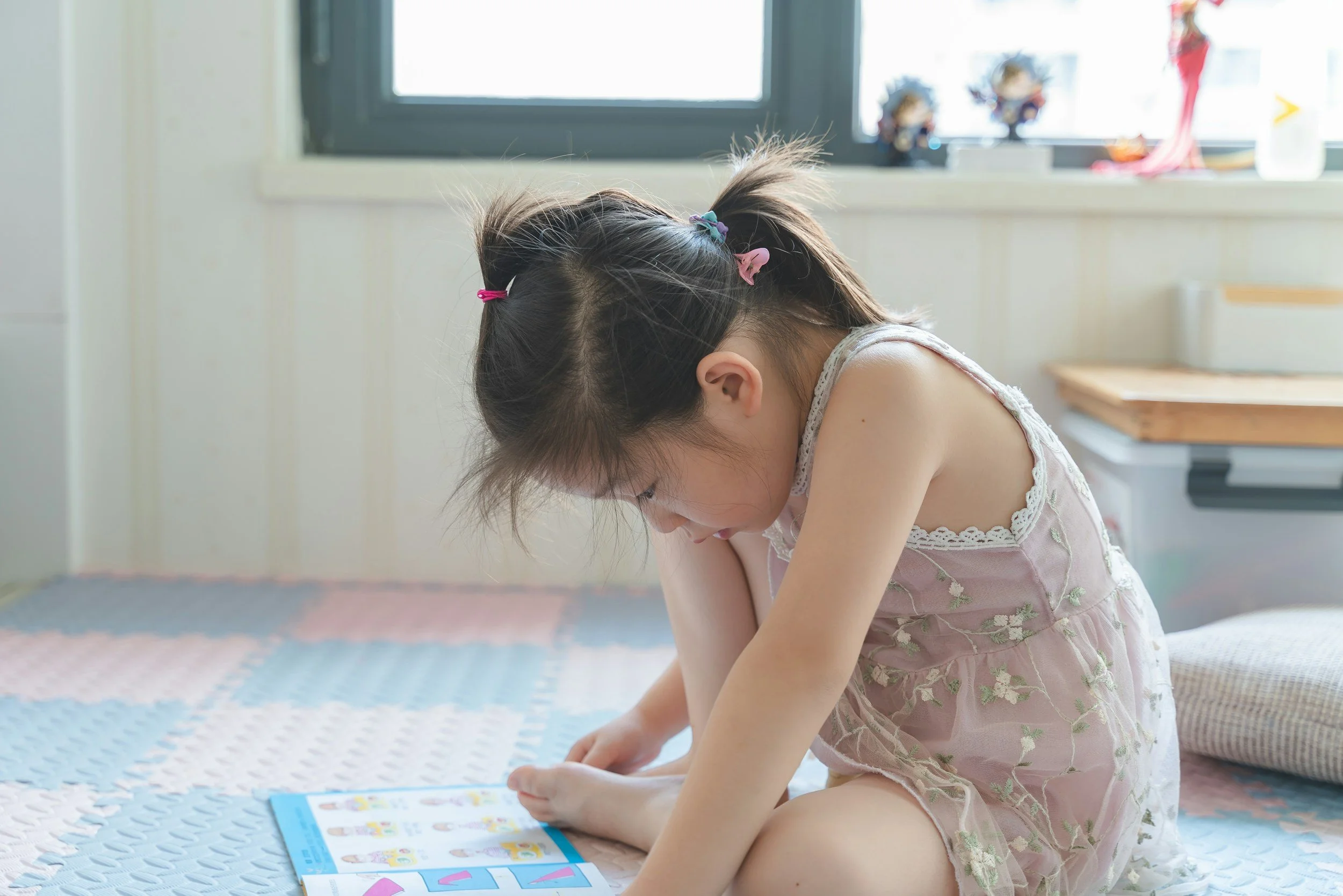 Young girl sitting on bed, looking at a colorful book, in a well-lit room with a window and various toys in the background.
