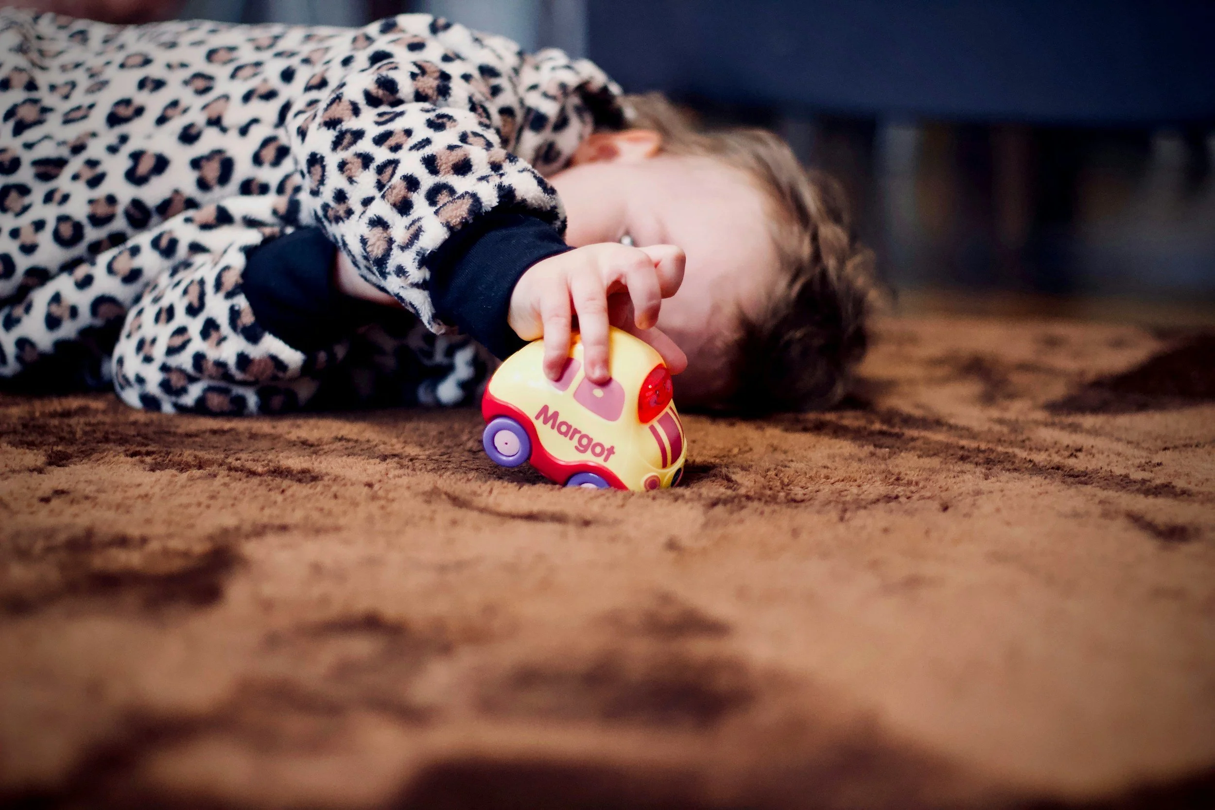 A young child lying on a brown carpet, holding a small yellow and red toy car labeled "Margot" with purple wheels, wearing a leopard print pajamas with black cuffs.
