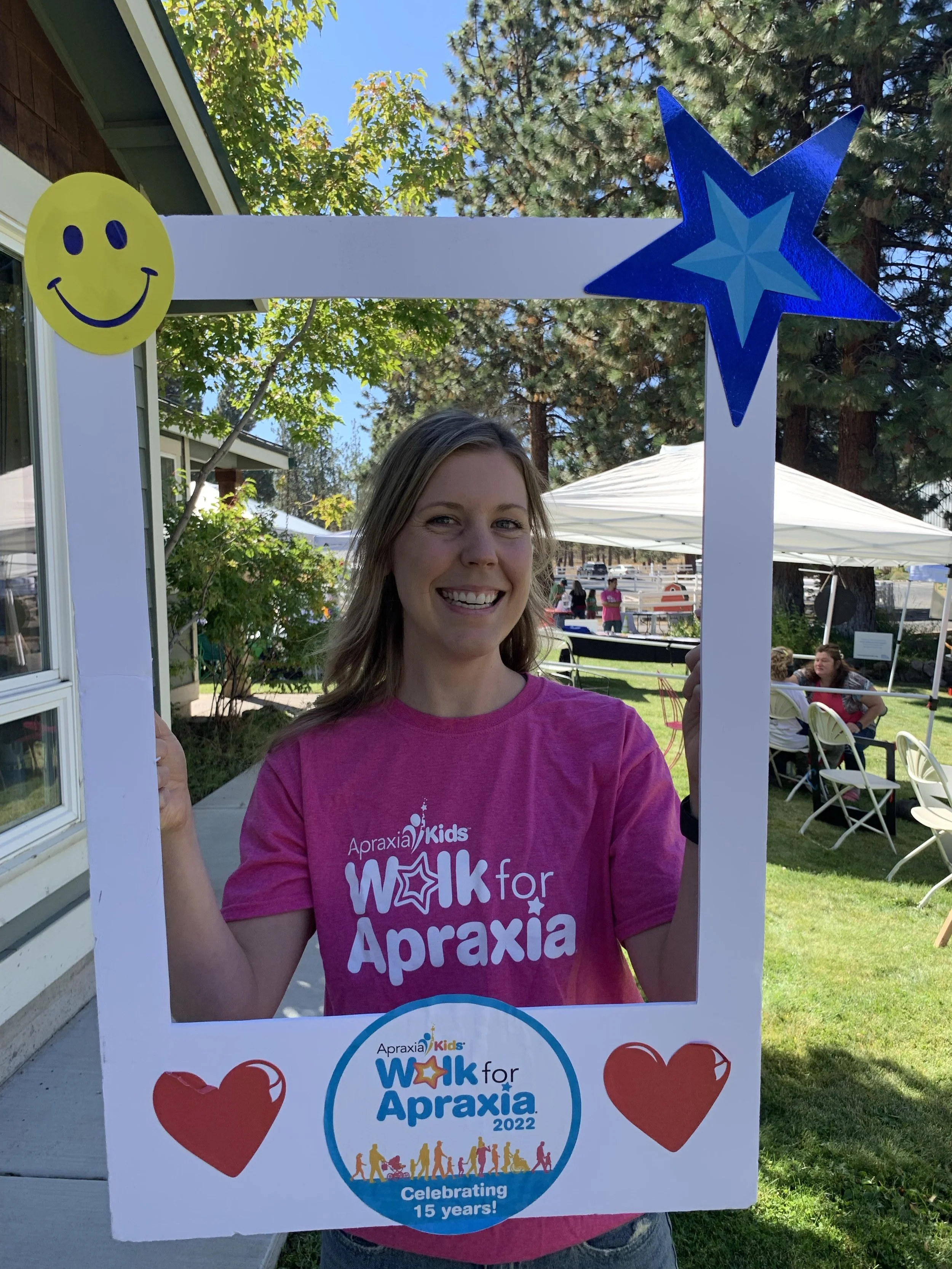 A woman smiling and holding a large photo frame with the logo for the 2022 Walk for Apraxia event, decorated with a yellow smiley face sticker, a blue star sticker, and two red heart stickers. The background shows people outdoors under tents, trees, and a grassy area.