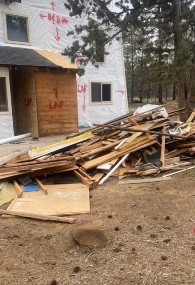 View of a house under construction with white house wrap and windows, and a pile of wooden debris and construction materials in front of it in a wooded area.