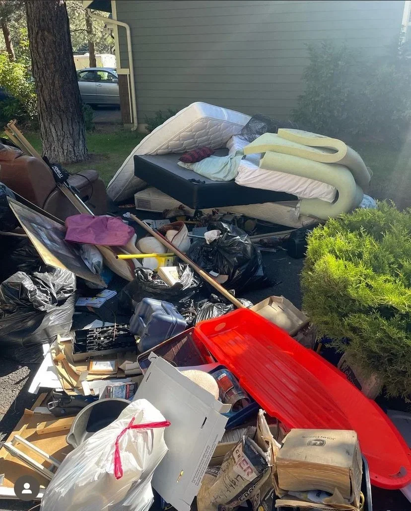 Pile of miscellaneous trash including mattresses, a red sled, boxes, plastic bags, and various household items scattered outside.