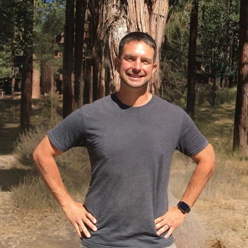 A man smiling outdoors in a forested area with tall trees, wearing a gray t-shirt and a wristwatch.