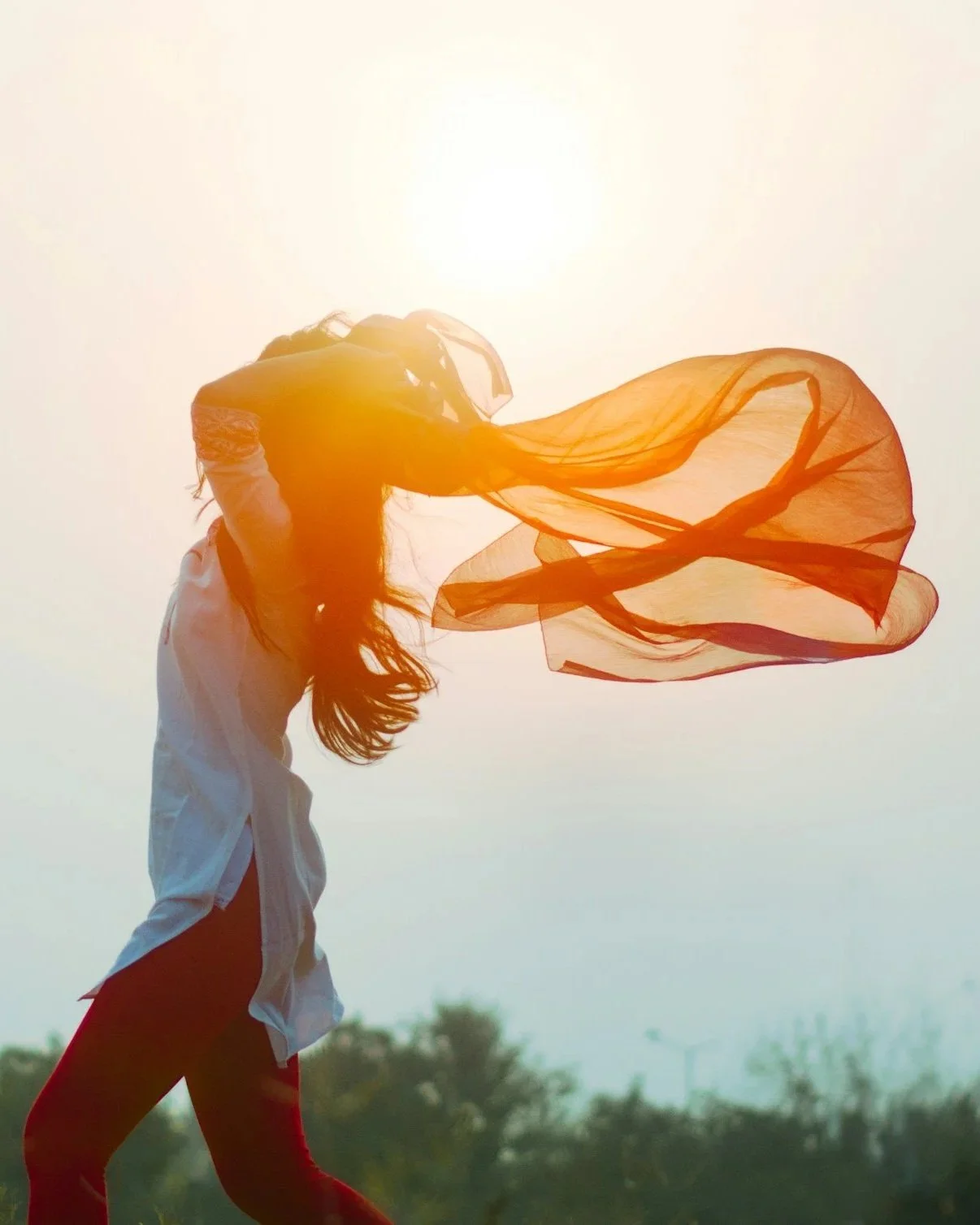 A woman standing outdoors in a light-colored shirt and red pants, holding a flowing, sheer orange scarf at sunset with the sun behind her.