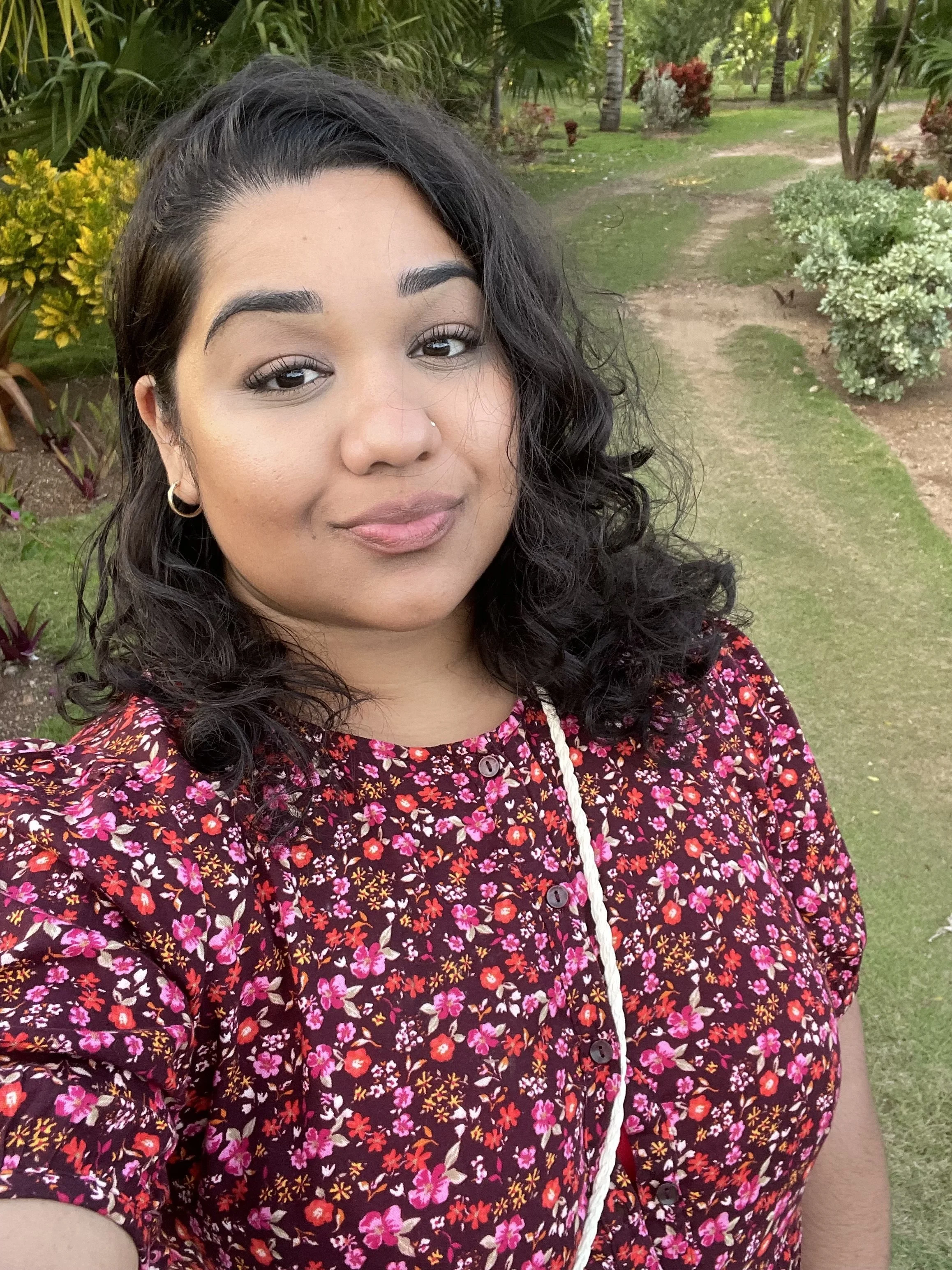 A woman with dark curly hair, wearing a floral dress and hoop earrings, standing outdoors in a garden with various plants and trees.