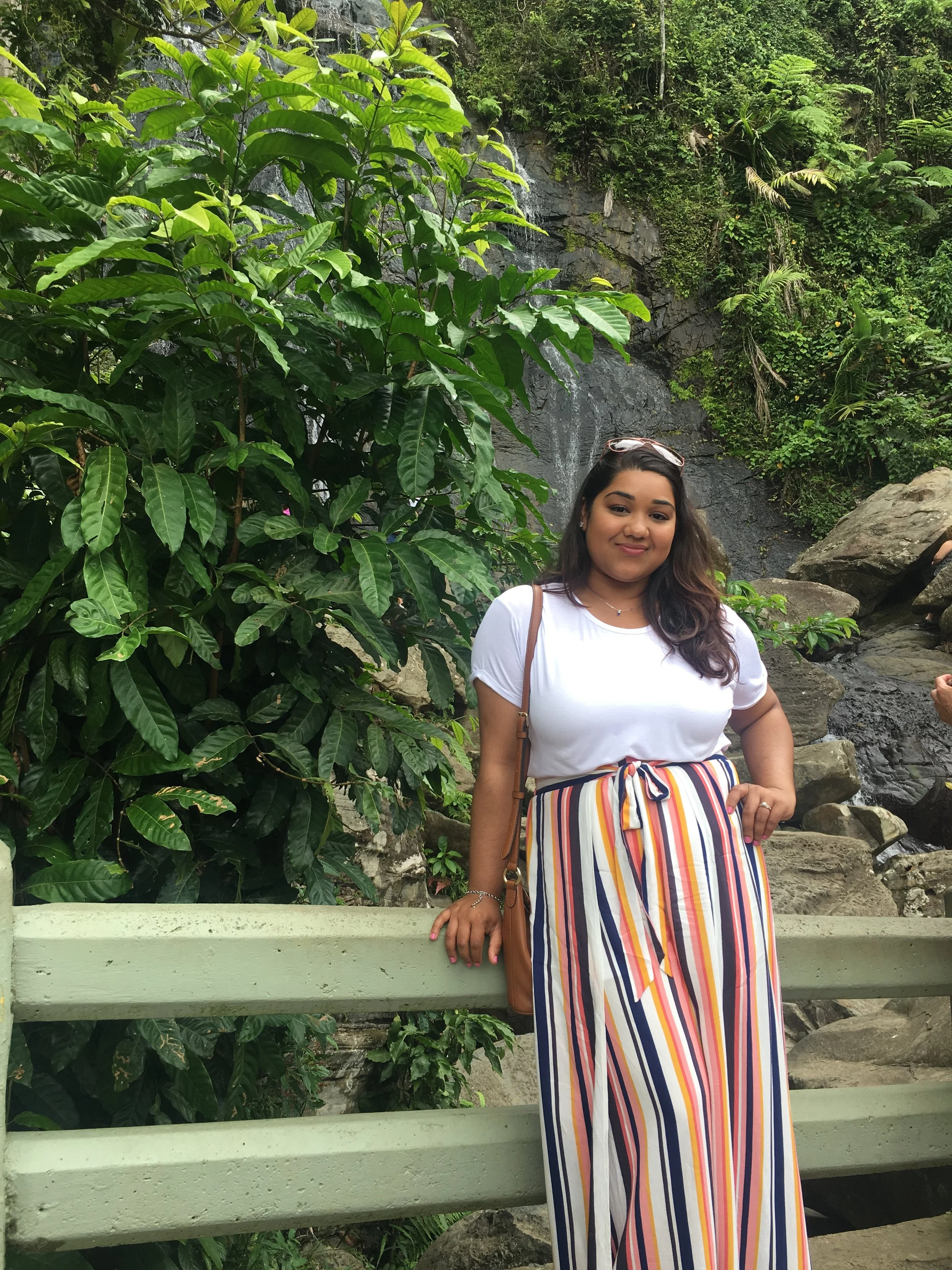 A young woman standing outdoors in front of a lush green landscape with a small waterfall, rocks, trees, and large leafy plants, wearing a white t-shirt, striped skirt, and sunglasses on her head.