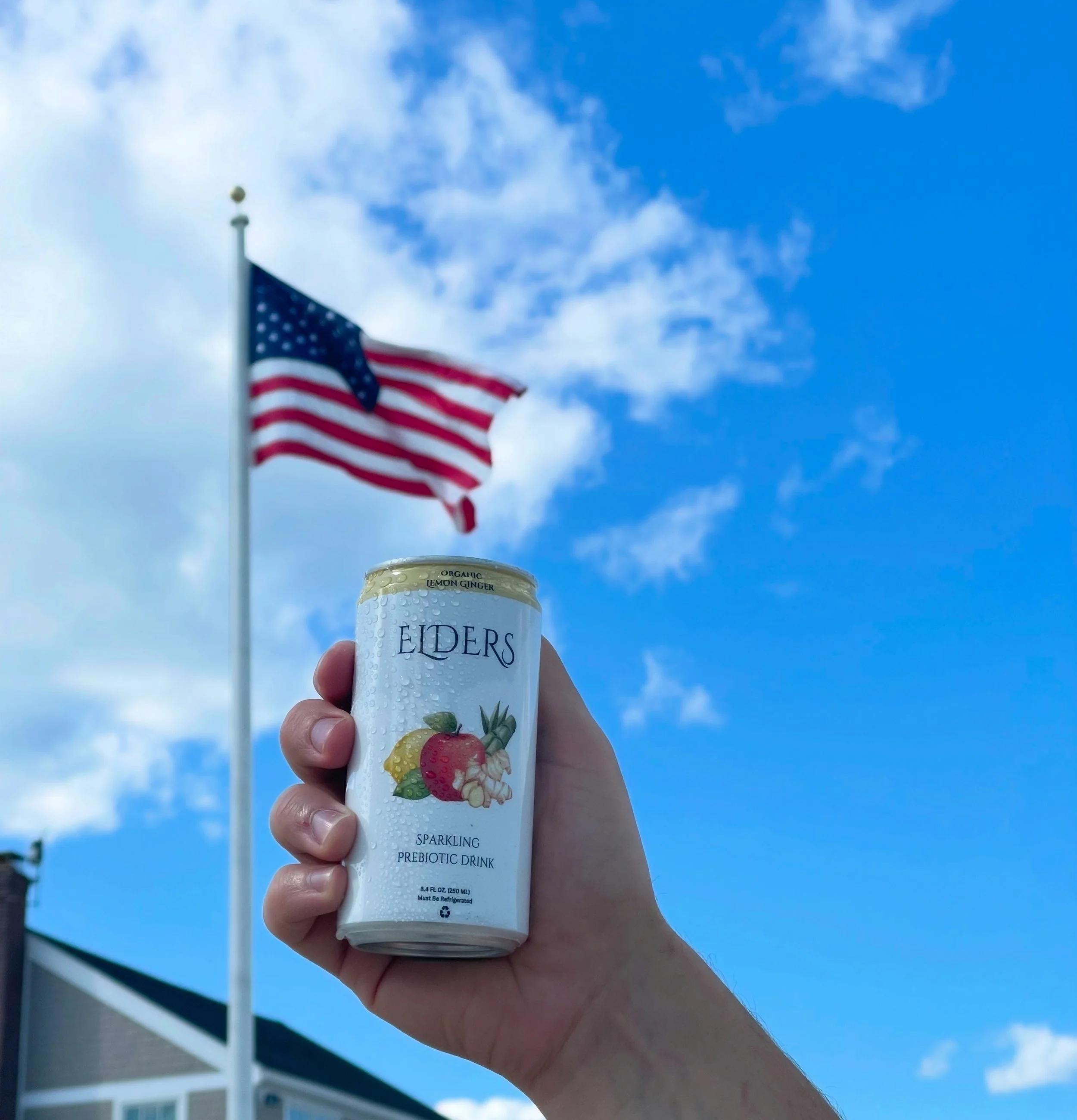 A hand holding a can of Eiders, a sparkling prebiotic drink, with an American flag in the background against a blue sky with scattered clouds.