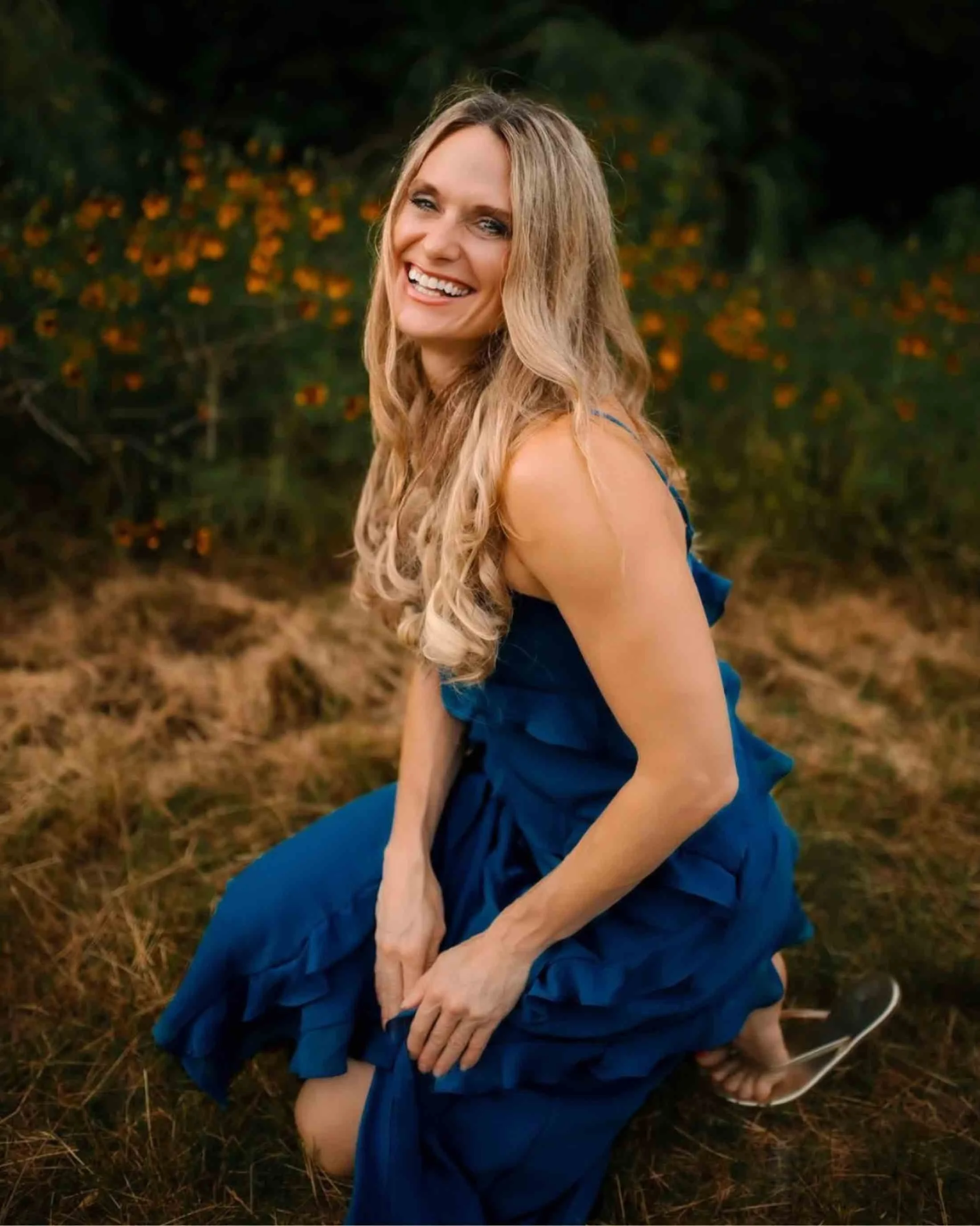 A woman with long, curly blonde hair smiling while kneeling outdoors on a grassy area, wearing a blue dress, with orange flowers and greenery in the background.