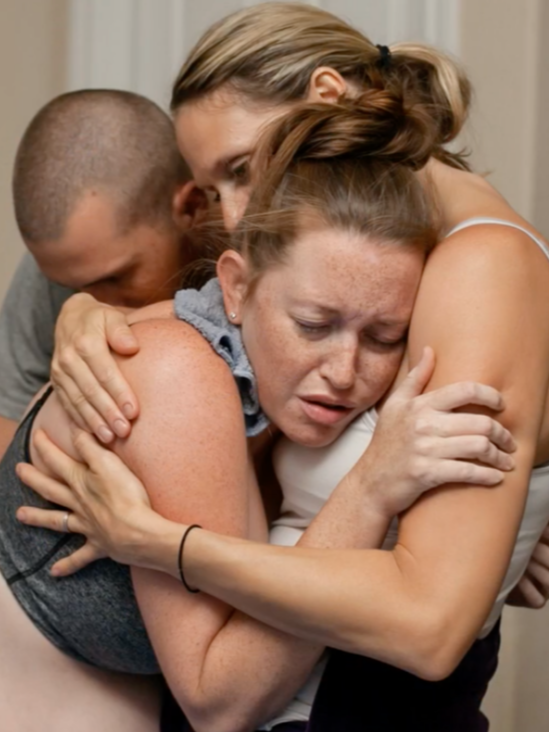 Three people hugging intimately with emotional expressions in a close embrace.