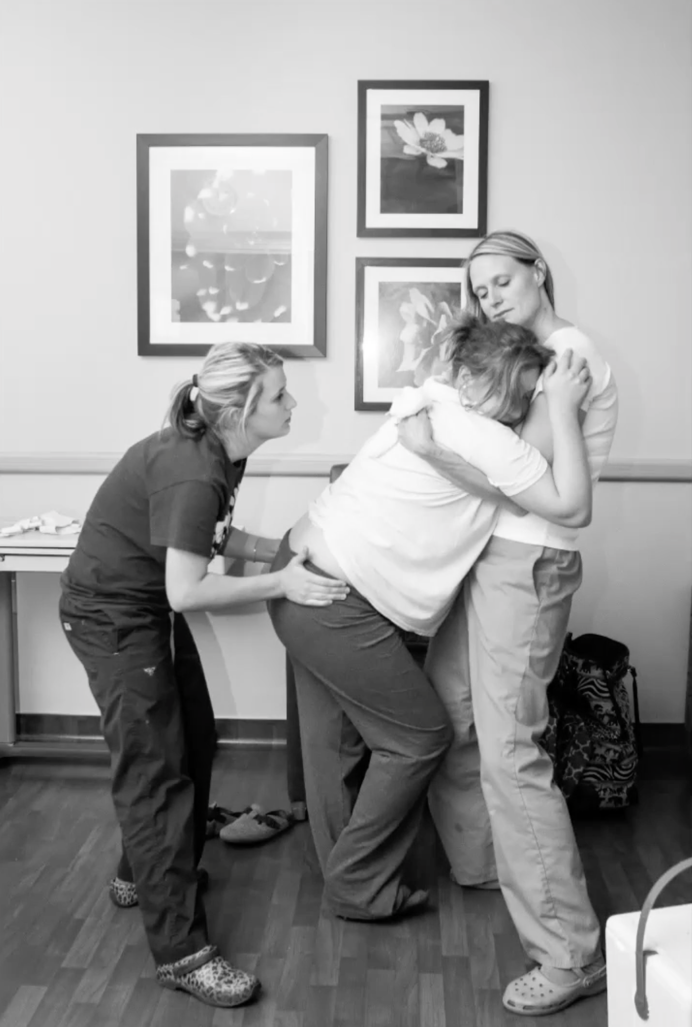 A woman appears to be in active labor with a healthcare professional supporting her, while another person holds her in a comforting embrace in a hospital or clinic room.