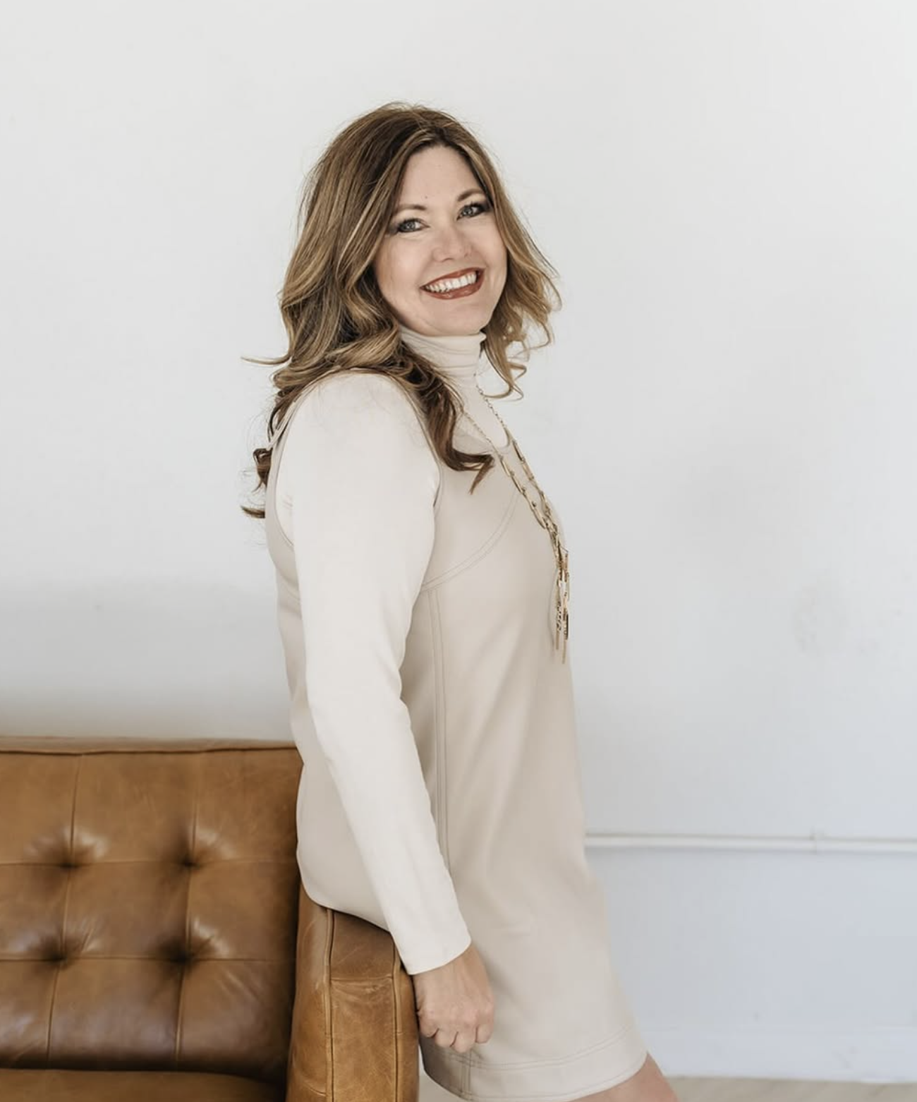 A woman with wavy brown hair, wearing a light-colored dress and a long chain necklace, smiling and leaning on a brown leather couch against a plain white wall.