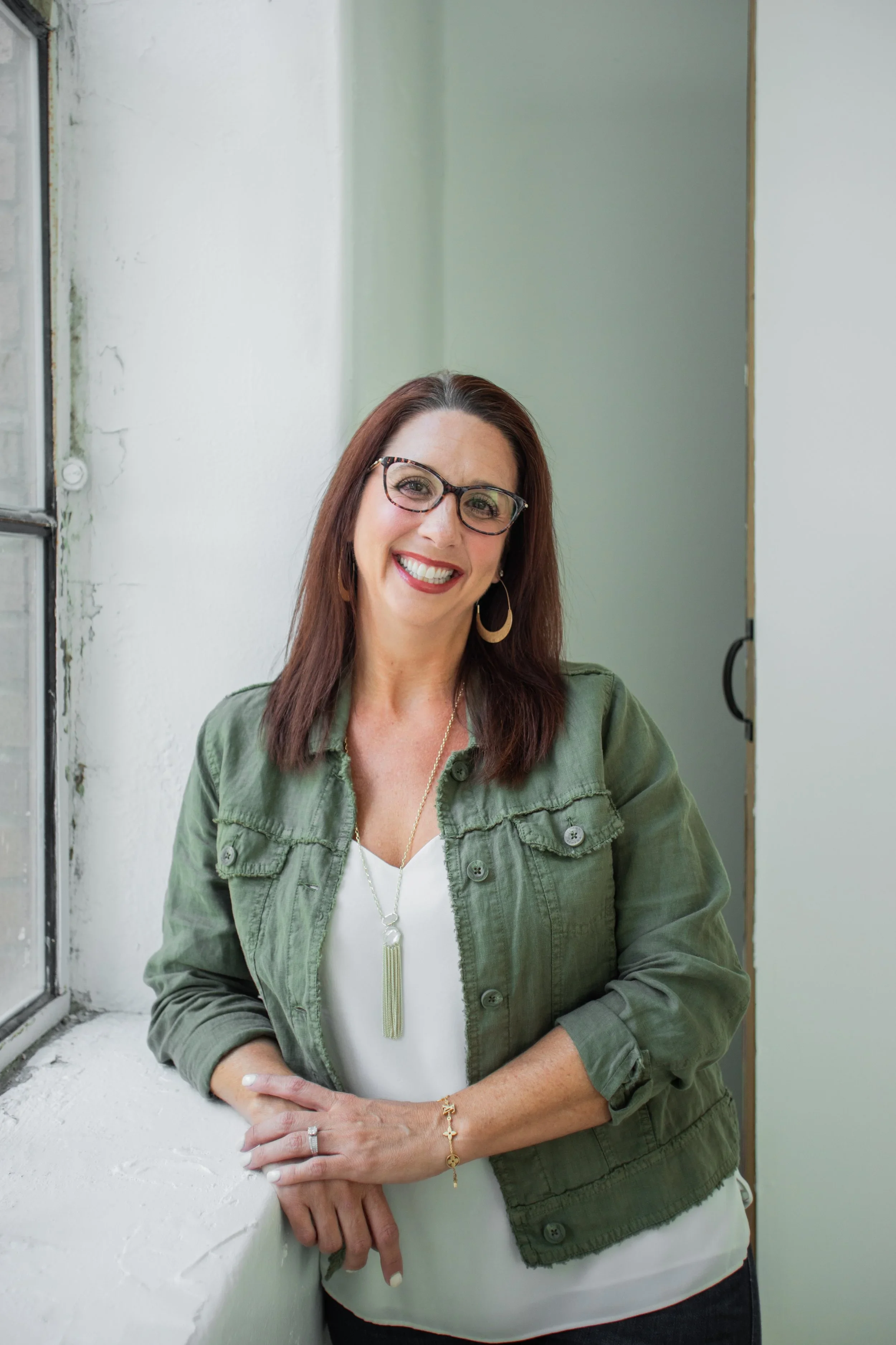 A smiling woman with brown hair, glasses, and hoop earrings, wearing a green jacket over a white shirt, standing by a window in a bright room.