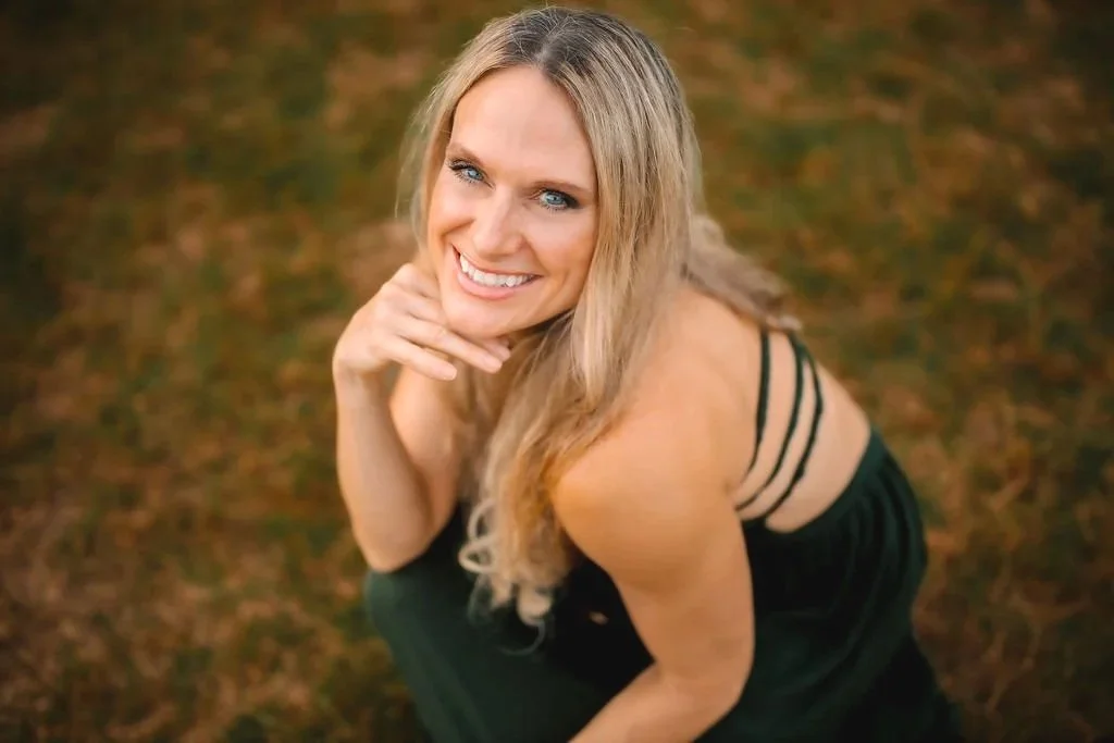 A woman with long blonde hair smiling and crouching outdoors on a grassy area during daytime.