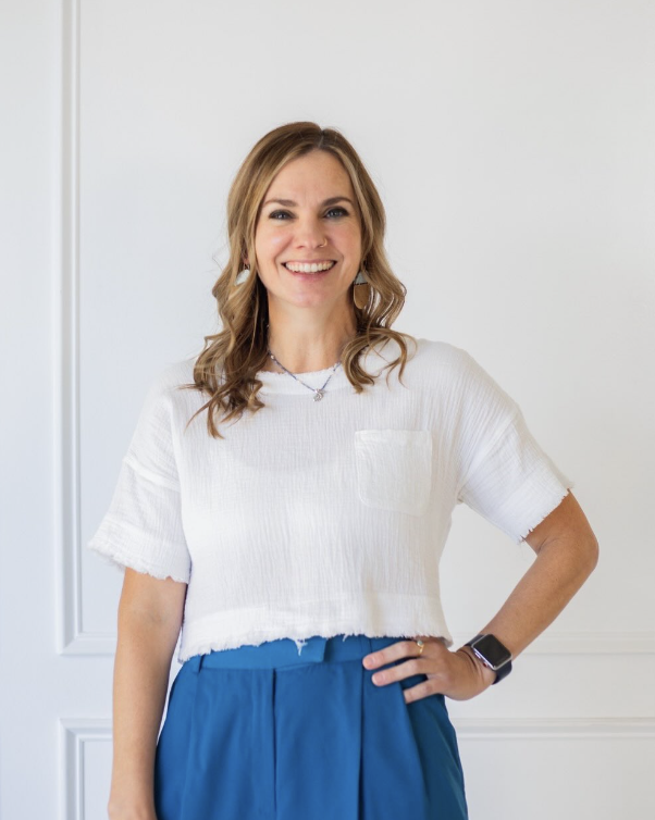 A smiling woman with wavy, shoulder-length brown hair, wearing a white short-sleeve top and blue pants, standing against a plain white wall.