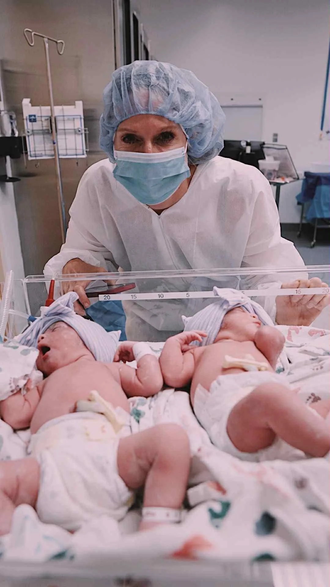 A nurse in scrubs, wearing a surgical mask, hair cover, and gloves, stands behind a clear incubator with newborn twins inside, in a hospital nursery.