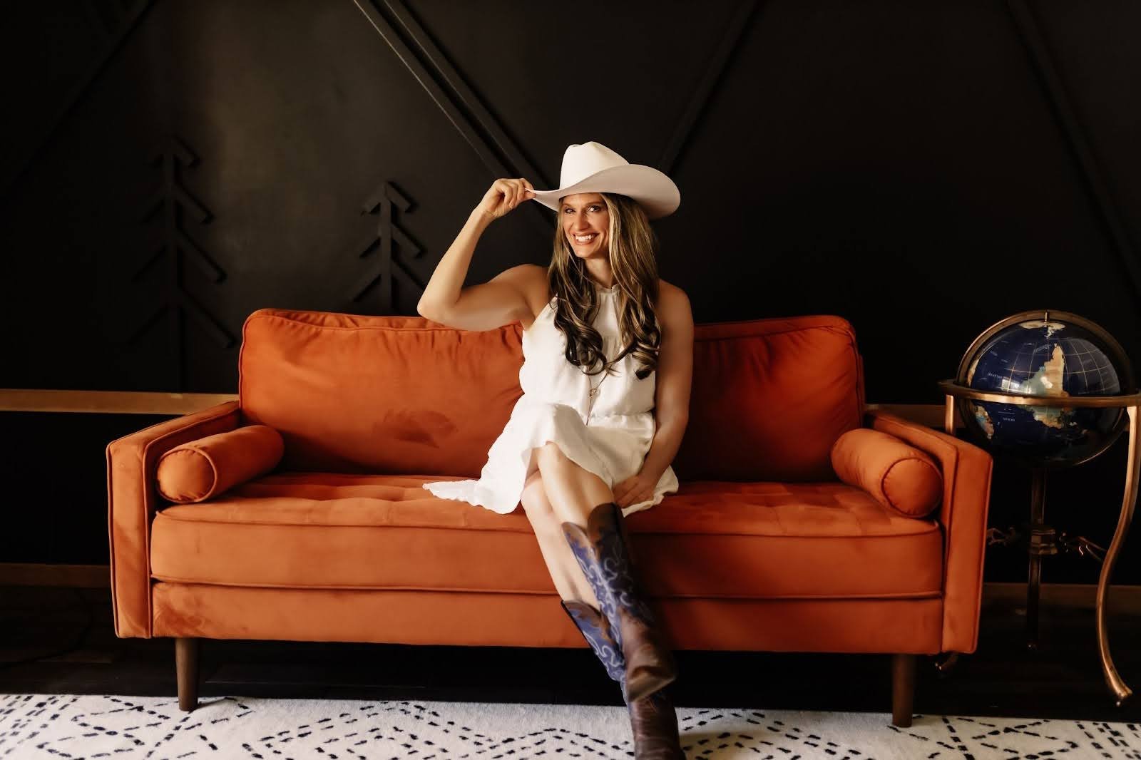 Woman in a white dress and cowboy hat sitting on an orange sofa, smiling, with a globe on a stand beside her.
