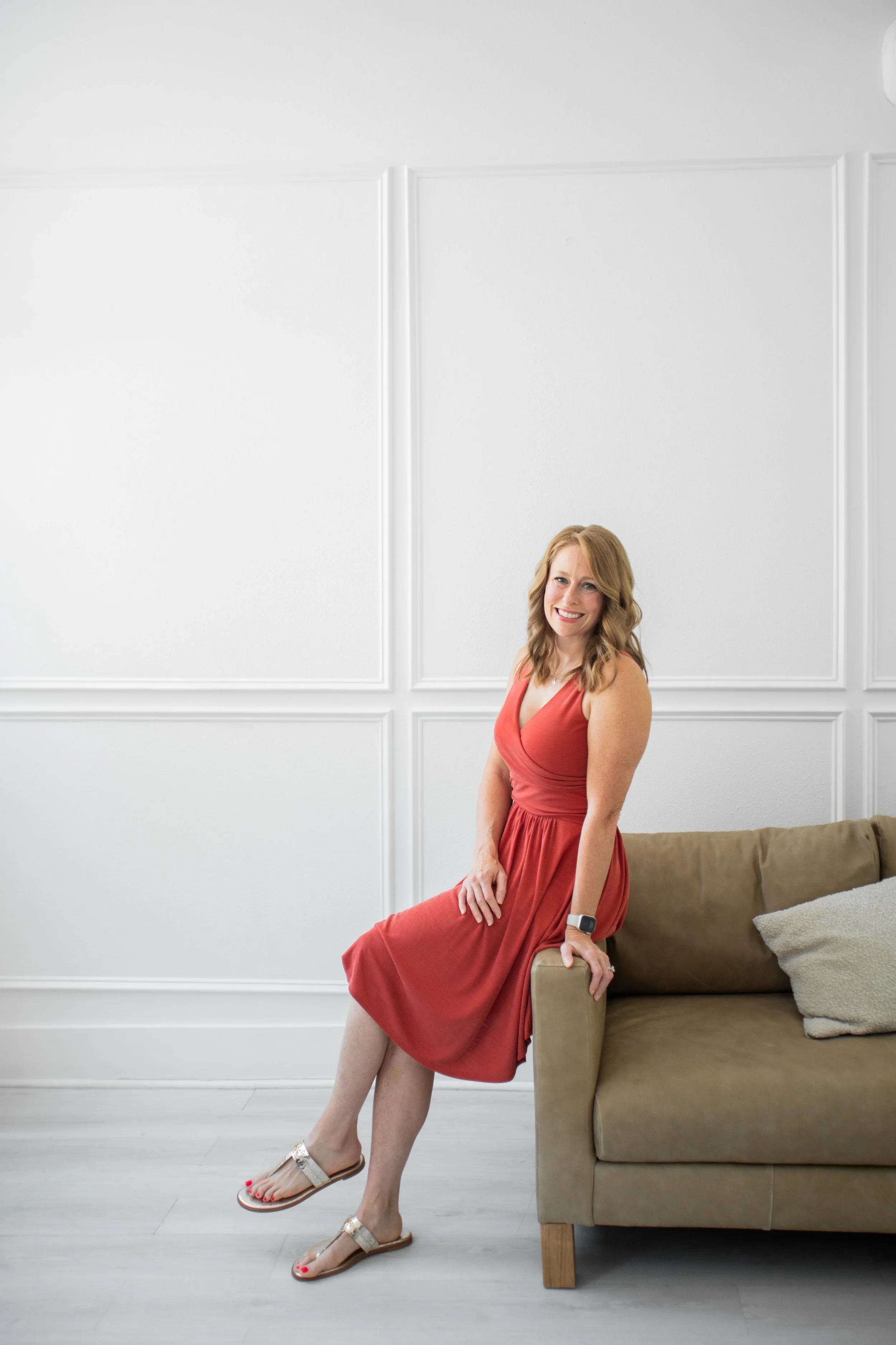 Woman in a red dress sitting on the arm of a beige sofa in a minimal white room.